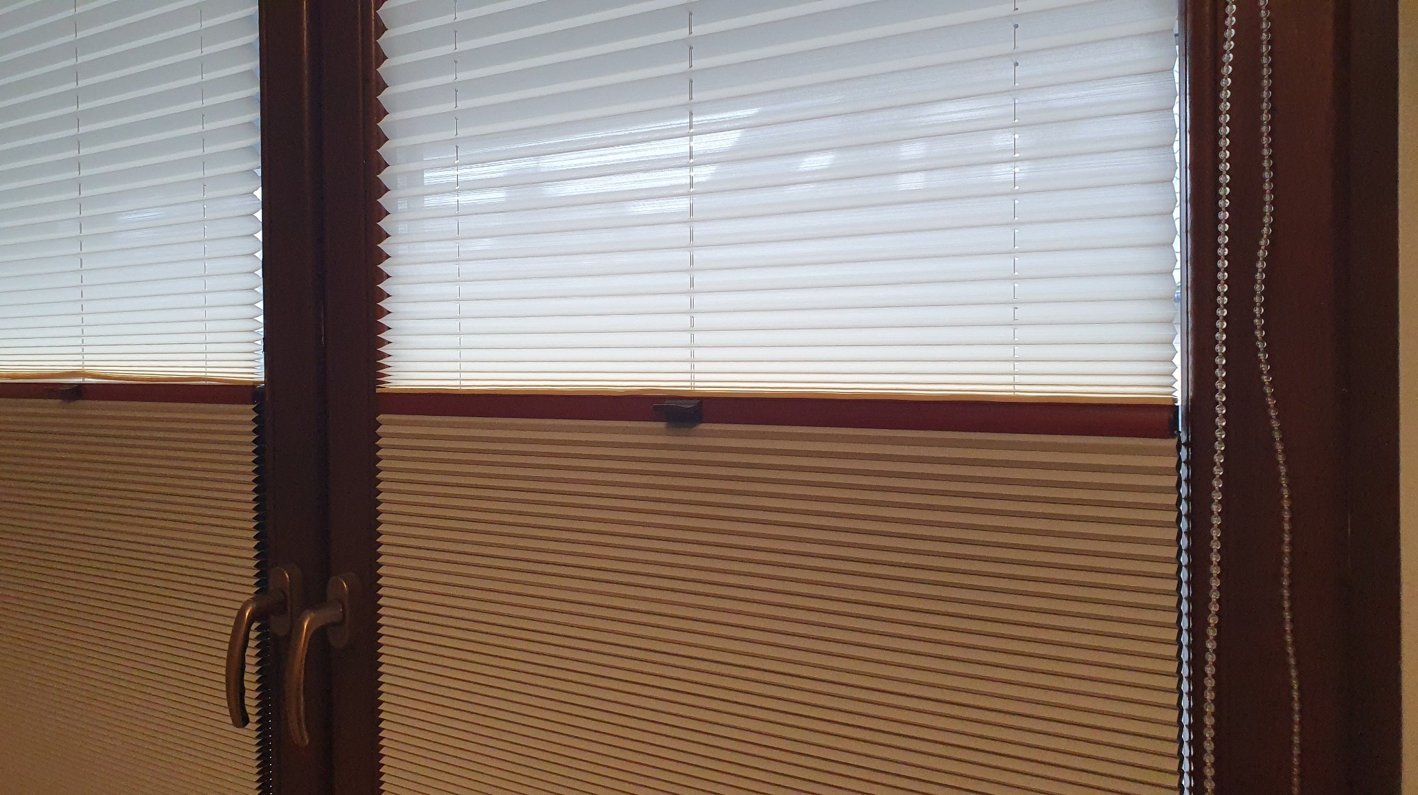 Beżowo-białe, podwójne, harmonijkowe osłony okienne z mechanizmem łańcuszkowym i widocznymi uchwytami, zamontowane w oknie z ciemnego drewna.