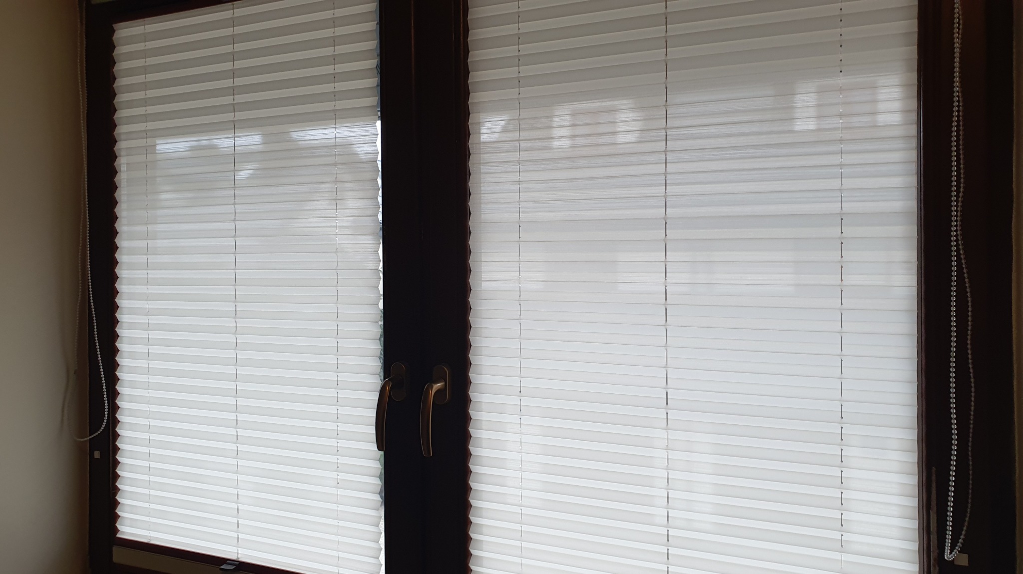 Białe, harmonijkowe osłony okienne zamontowane w ciemnej ramie okiennej. Widoczny sznurek sterujący po prawej stronie.