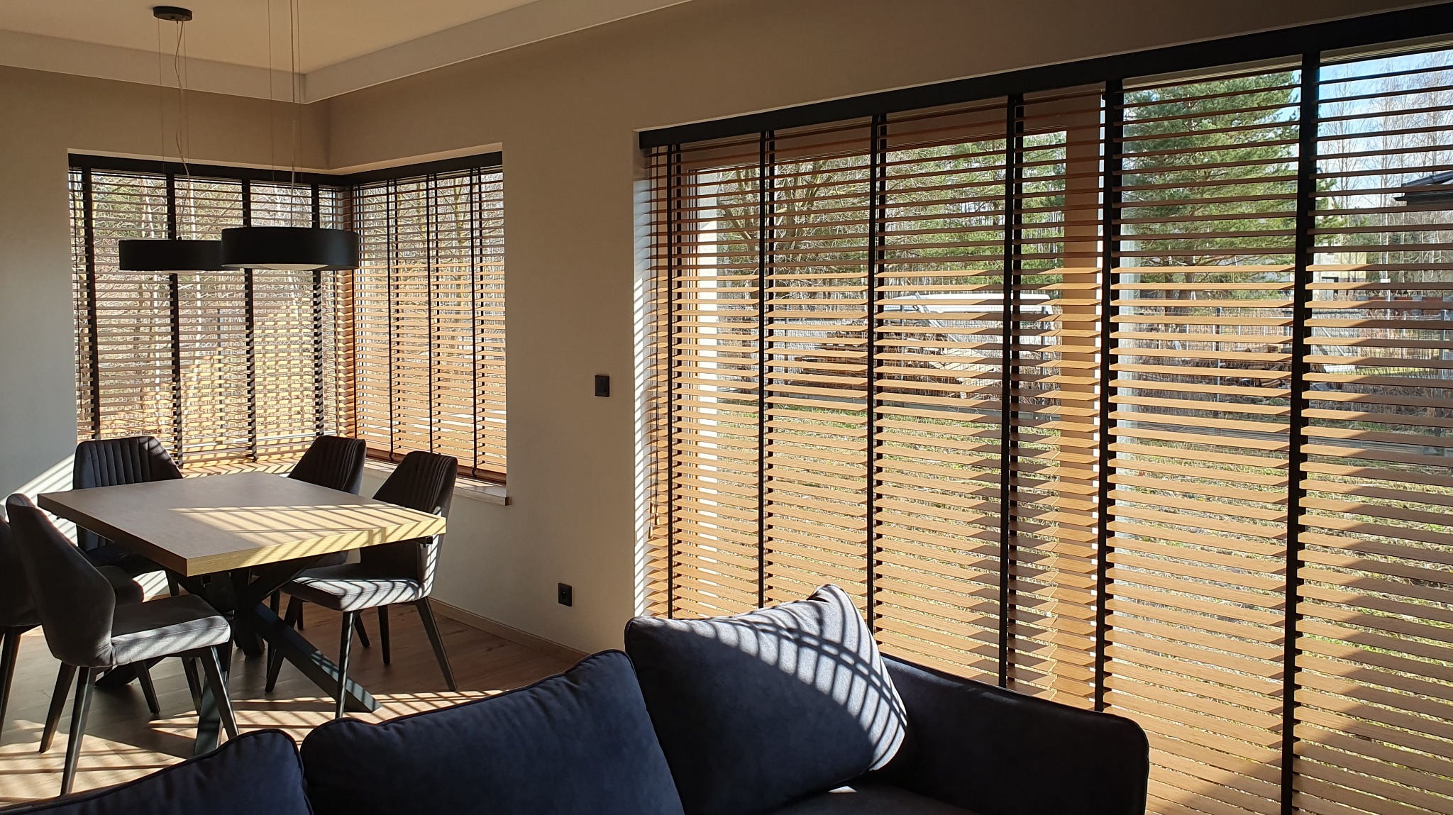 Jasne drewniane żaluzje z czarnymi elementami w nowoczesnym salonie. Promienie słoneczne wpadają przez okna, oświetlając stół i krzesła.