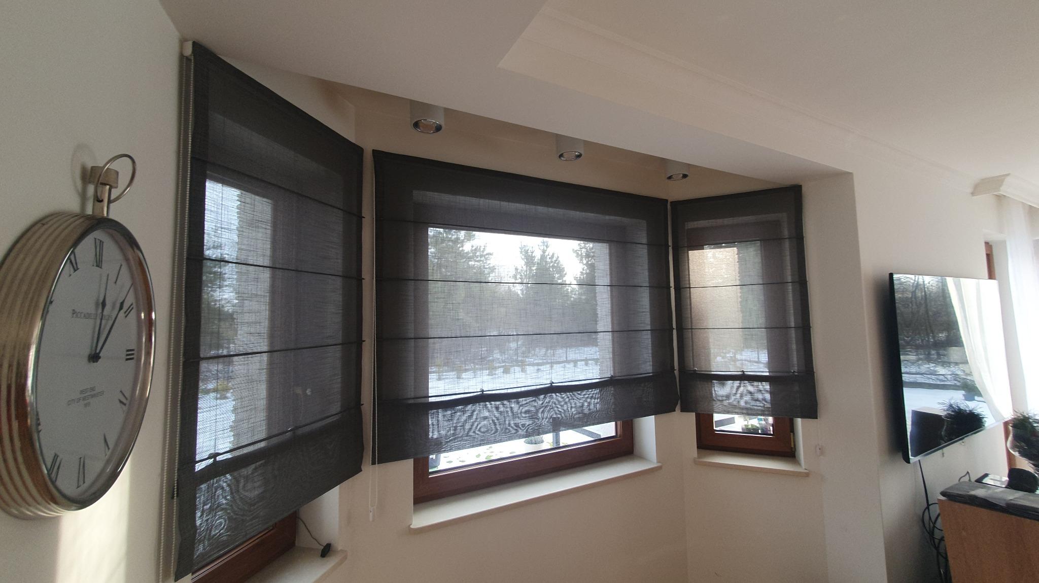Zestaw trzech grafitowych rolet rzymskich w oknach, widoczne delikatne poziome pasy i faktura materiału, zegar na ścianie po lewej stronie kadru.