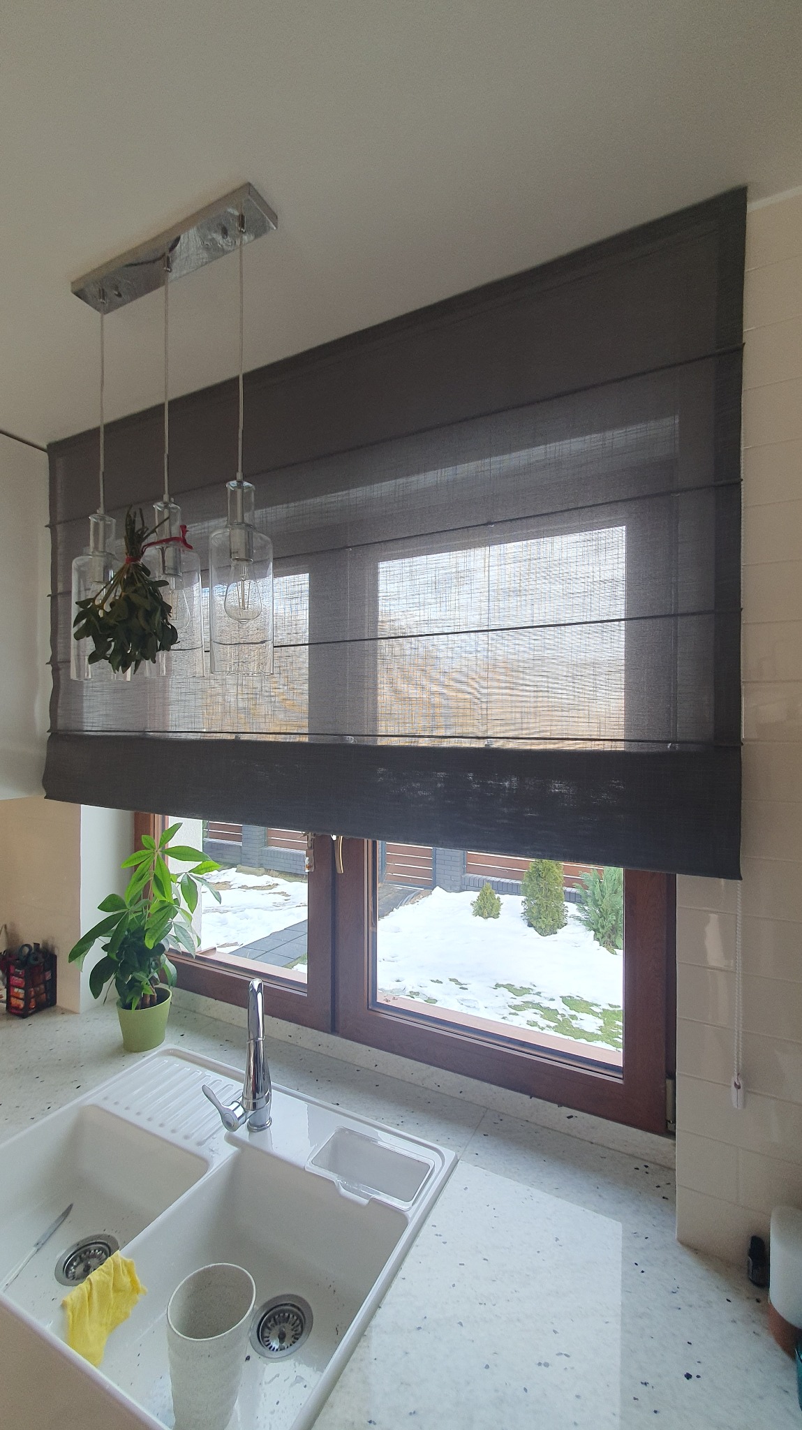 Szara roleta rzymska w oknie kuchni z widokiem na zaśnieżony ogród. Nad zlewem wiszą lampy z gałązkami jemioły. Neutralne światło dzienne.