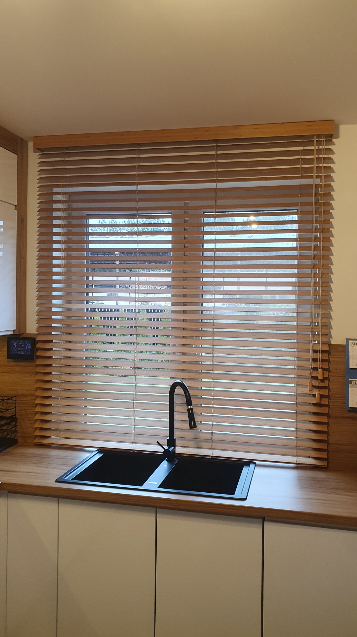 Kuchnia z oknem przesłoniętym drewnianymi żaluzjami w ciepłym odcieniu, czarny zlew dwukomorowy i bateria. Widok na zewnątrz przez lamele.