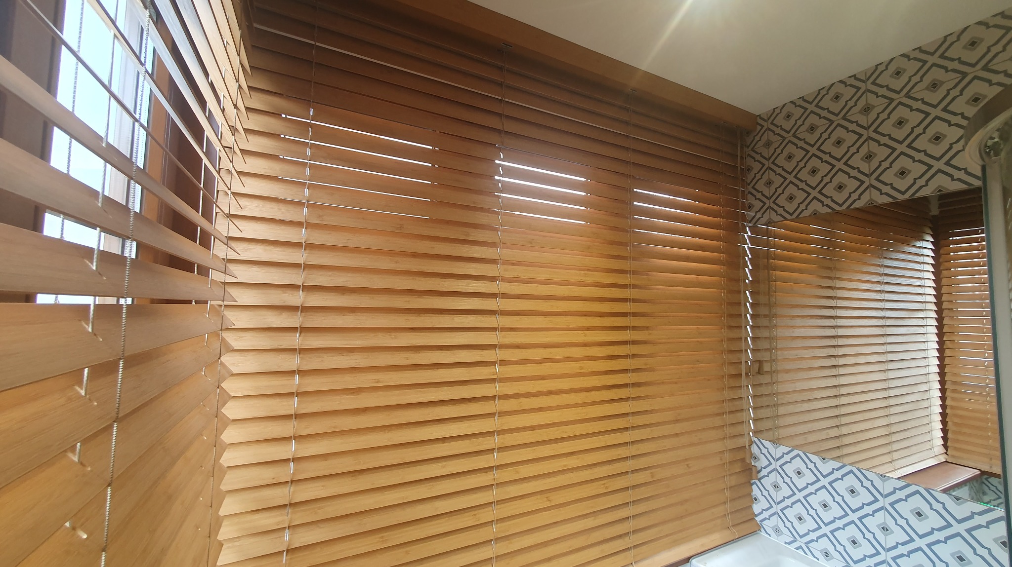 Jasne, drewniane żaluzje poziome zamontowane w łazience z wzorzystą tapetą. Częściowo odsłonięte, wpuszczają światło dzienne. Widok z bliska.