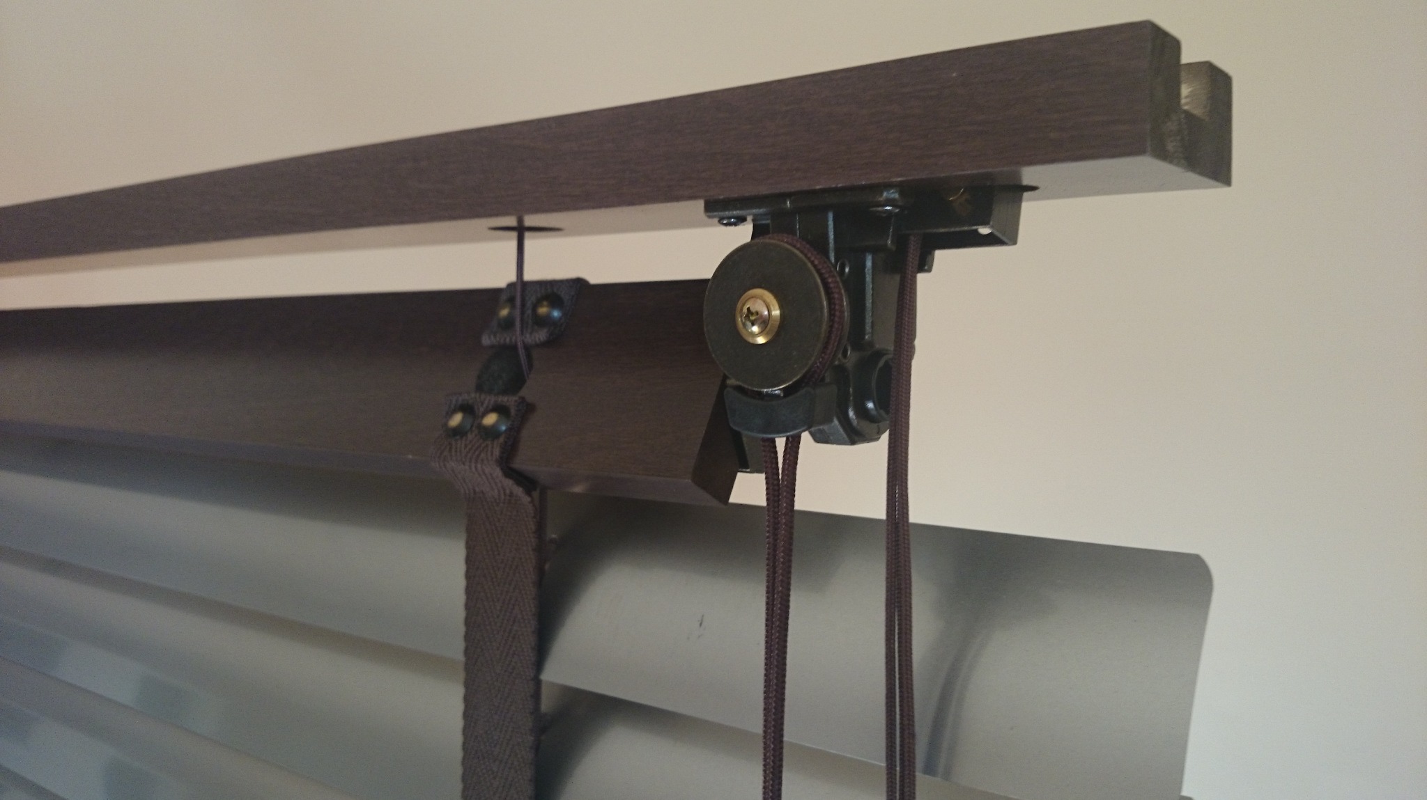 Fragment mechanizmu brązowych żaluzji z listwami w kolorze srebra, widoczne sznurki i elementy montażowe, całość na jasnym tle ściany.