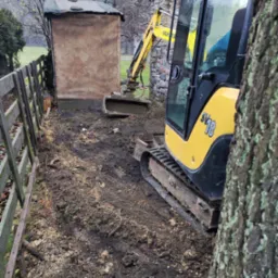 Żółta mini koparka Yanmar SV18 w trakcie prac ziemnych, widoczna obok drewnianego płotu i małej budki z kamienną elewacją, w tle drzewa bez liści.