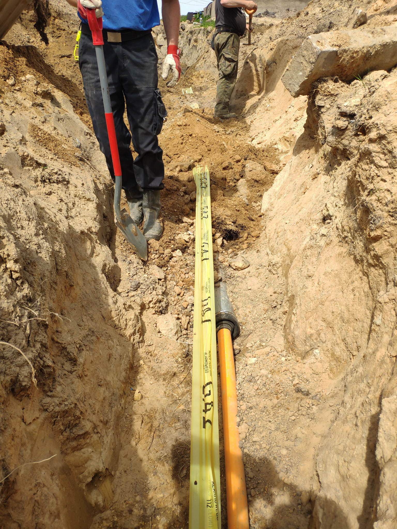 Wykop z ułożonymi rurami gazowymi i wodociągowymi, widoczni dwaj pracownicy z łopatami, jeden w czarnych spodniach i niebieskiej koszulce, drugi w spodniach moro, ziemia piaszczysta.