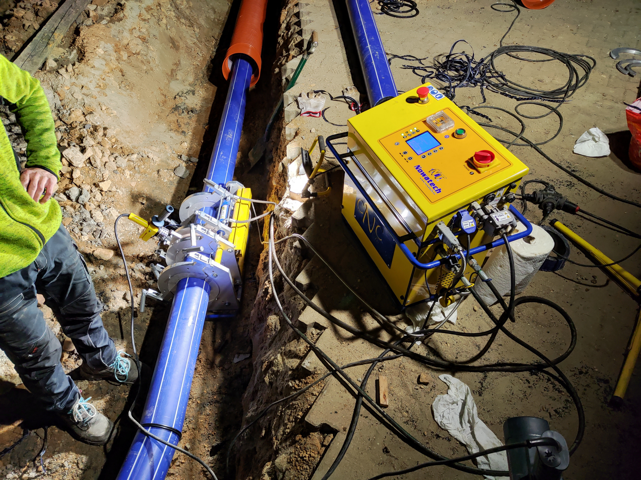 Wykop z ułożonymi rurami gazowymi z niebieskiego tworzywa, żółta maszyna do zgrzewania rur i fragment sylwetki pracownika w zielonej bluzie.