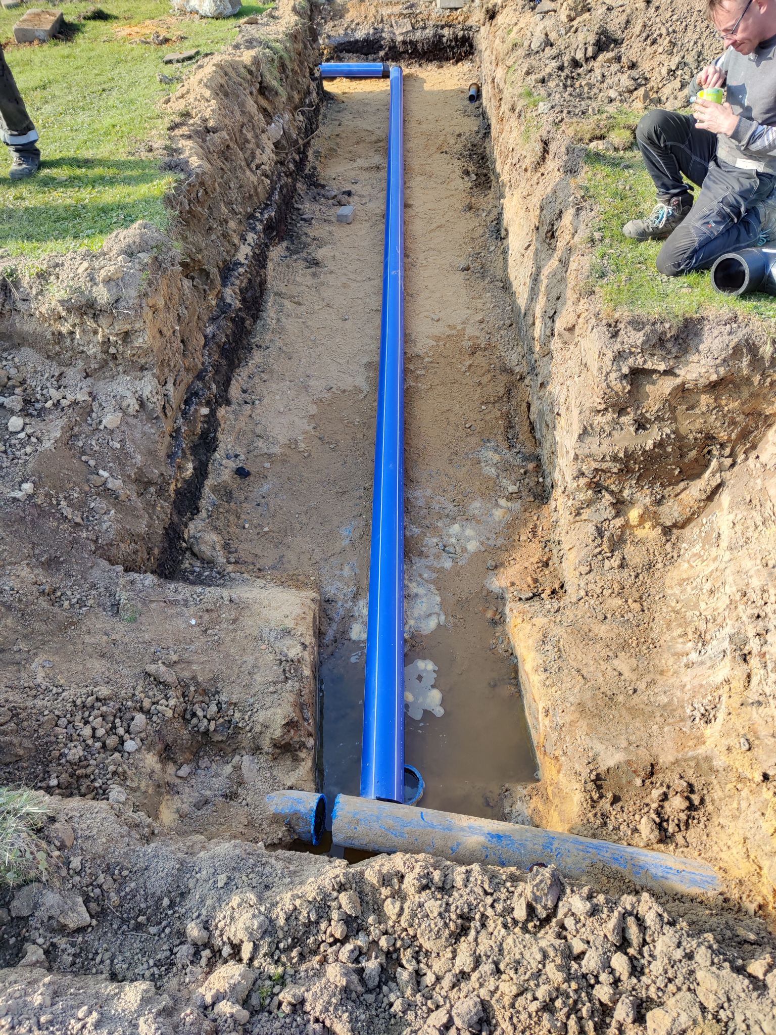 Wykop z ułożonym niebieskim rurociągiem wodnym, widoczny fragment instalatora w tle, woda w wykopie.