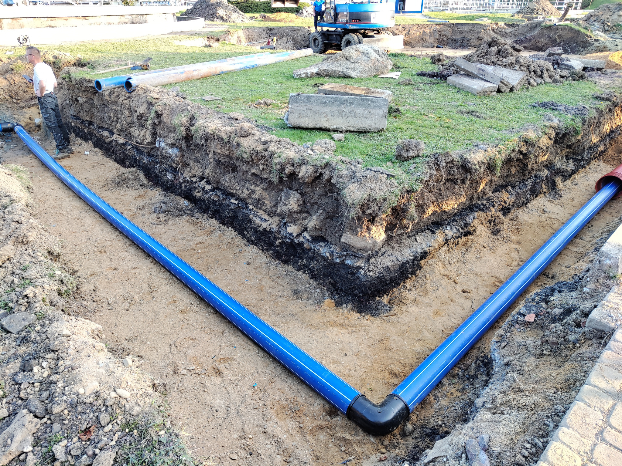 Niebieskie rury wodociągowe ułożone w wykopie, z widocznym łącznikiem kątowym, na tle ziemi i trawy, z pracującą koparką w oddali.