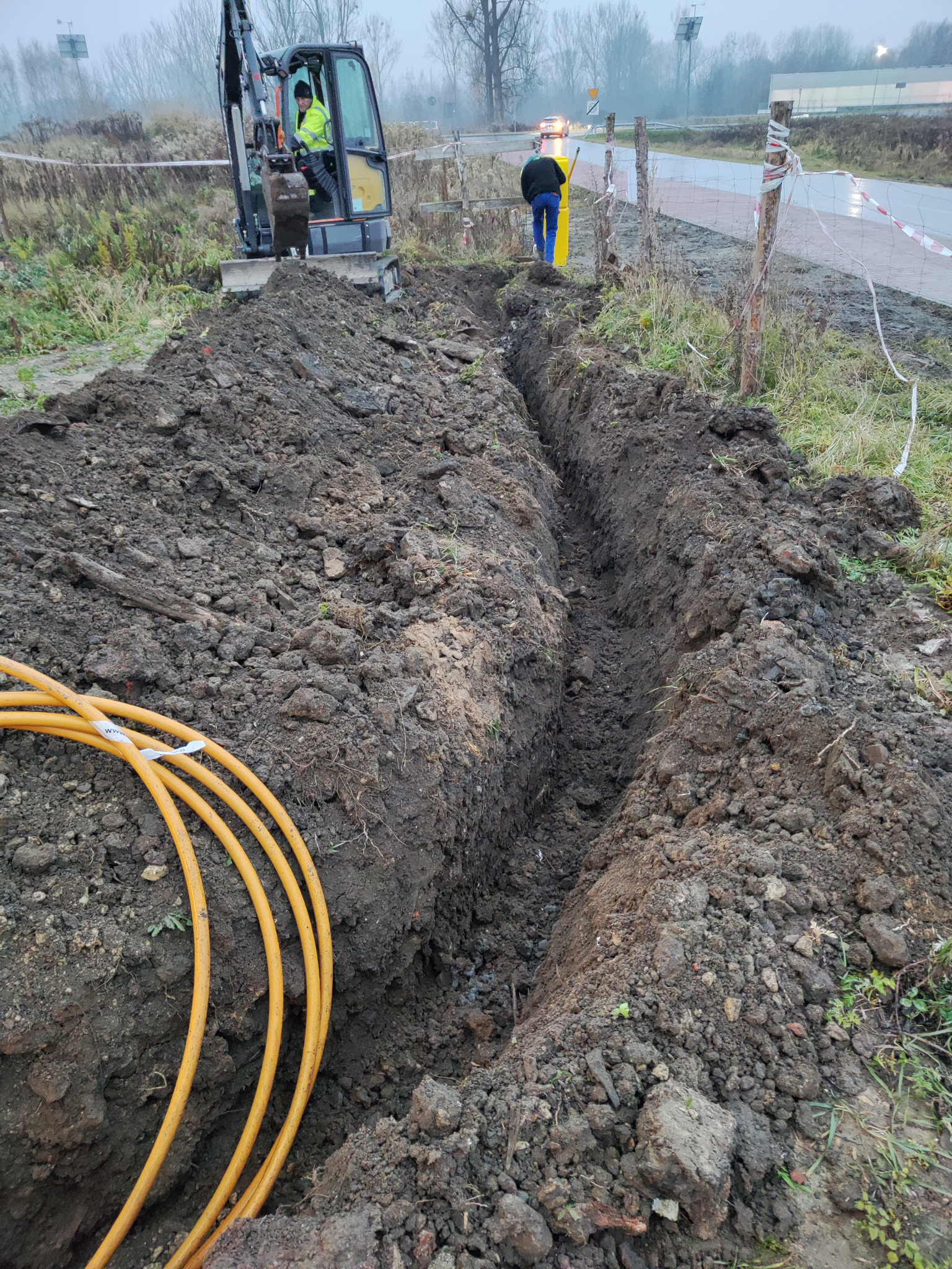 Wykopany rów w ziemi, widoczne żółte rury, pracownik w żółtej kamizelce obsługujący małą koparkę, zabezpieczenie wykopu taśmą ostrzegawczą.