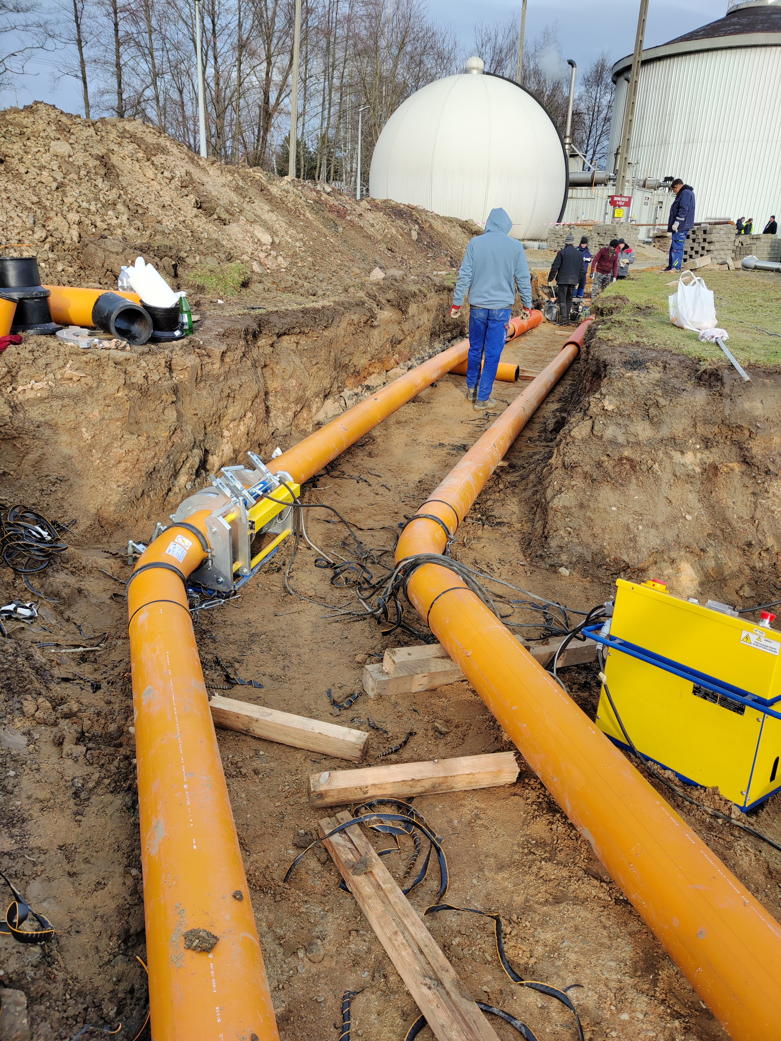 Pomarańczowe rury ułożone w wykopie, połączone złączką, w tle pracownicy i biały zbiornik, widoczny żółty agregat spawalniczy.