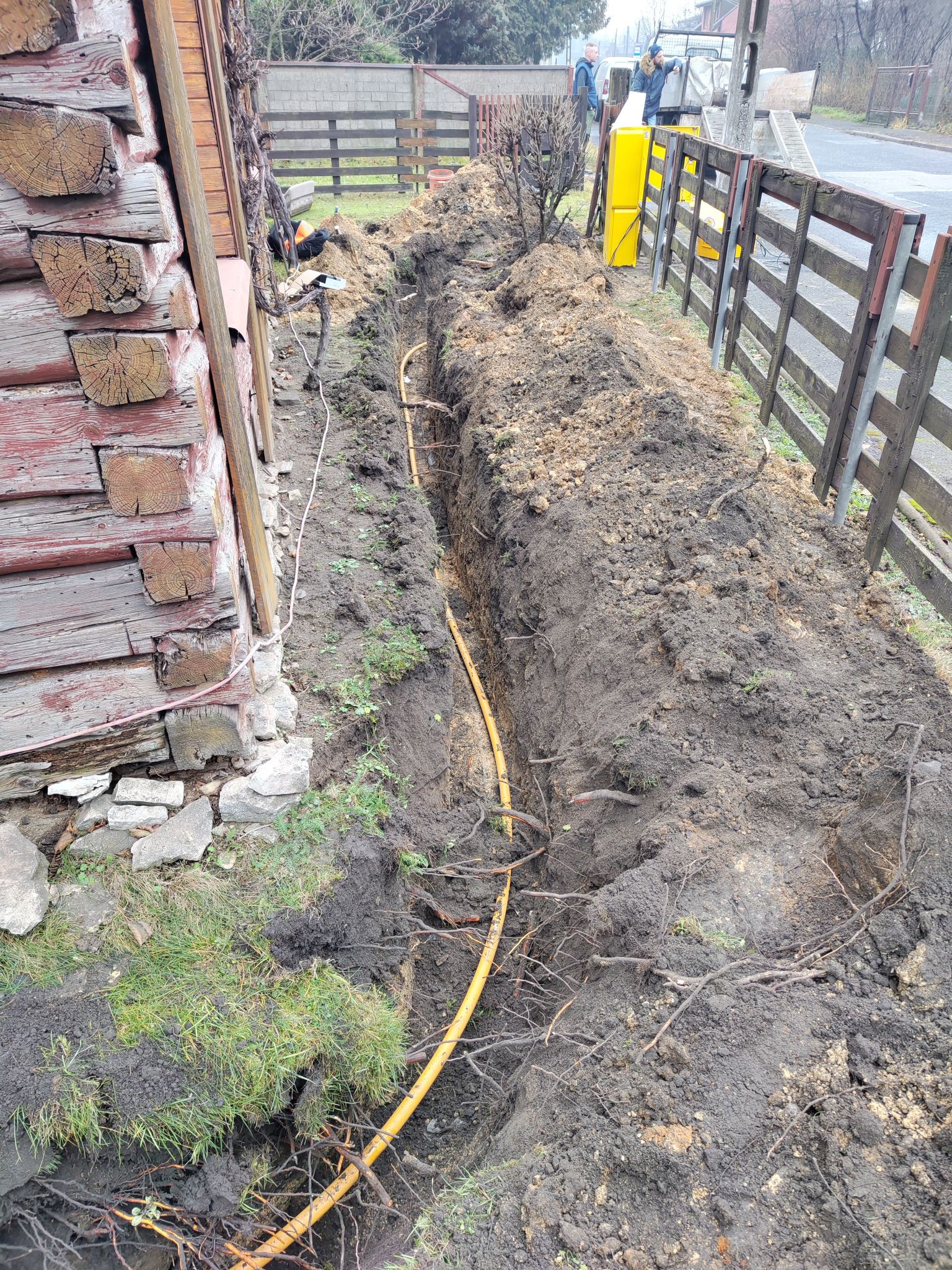 Żółty rurociąg ułożony w wykopie ziemnym wzdłuż ściany budynku z drewnianych bali, widoczny fragment ogrodzenia i pracownicy w tle.