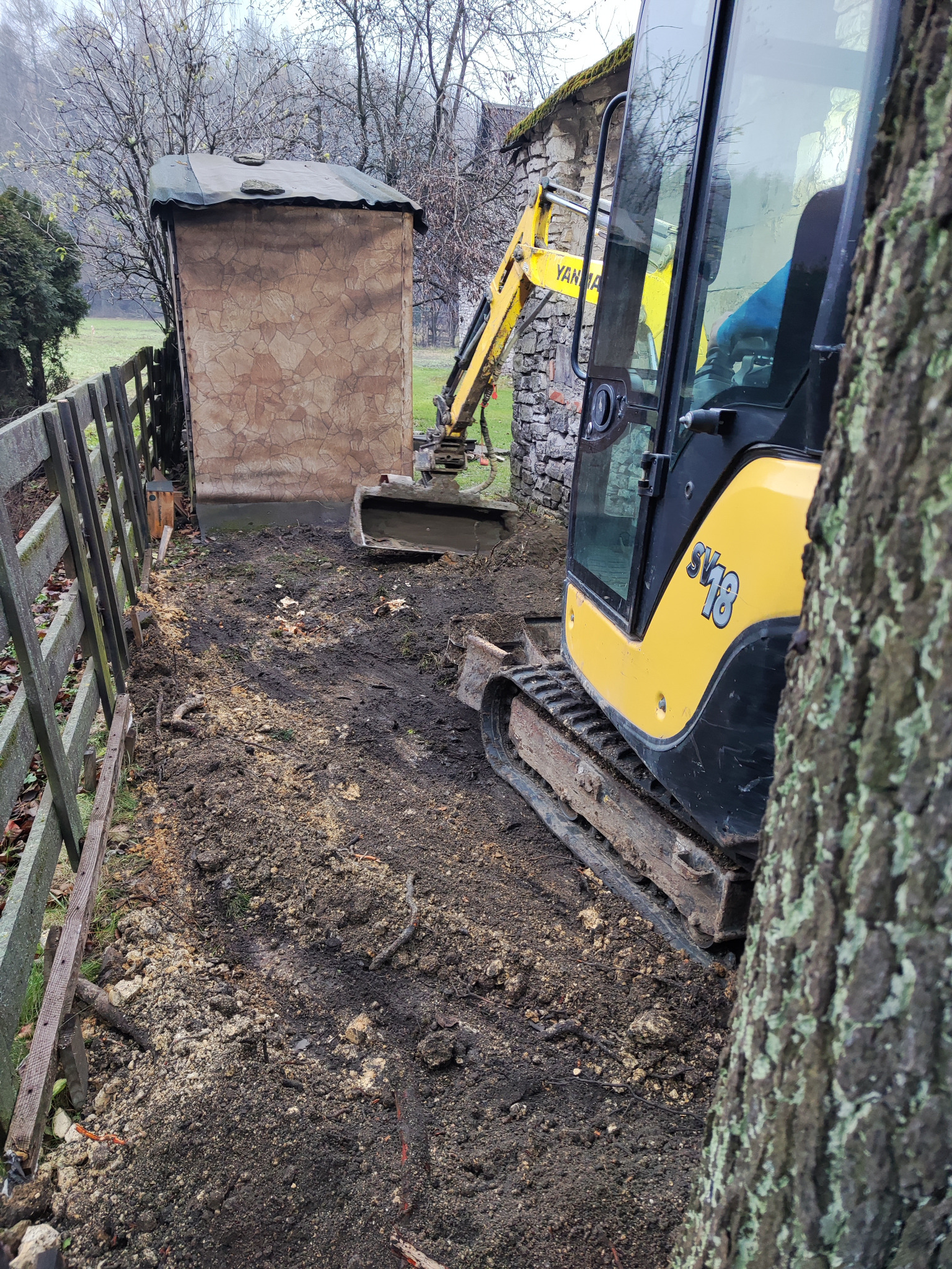 Żółta mini koparka Yanmar SV18 w trakcie prac ziemnych, widoczna obok drewnianego płotu i małej budki z kamienną elewacją, w tle drzewa bez liści.