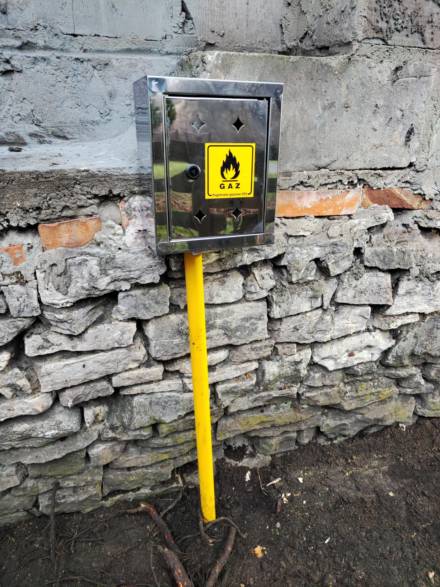 Metalowa skrzynka gazowa z żółtą rurą przyłączeniową, zamontowana na tle nierównej, kamiennej ściany z ceglanymi elementami.