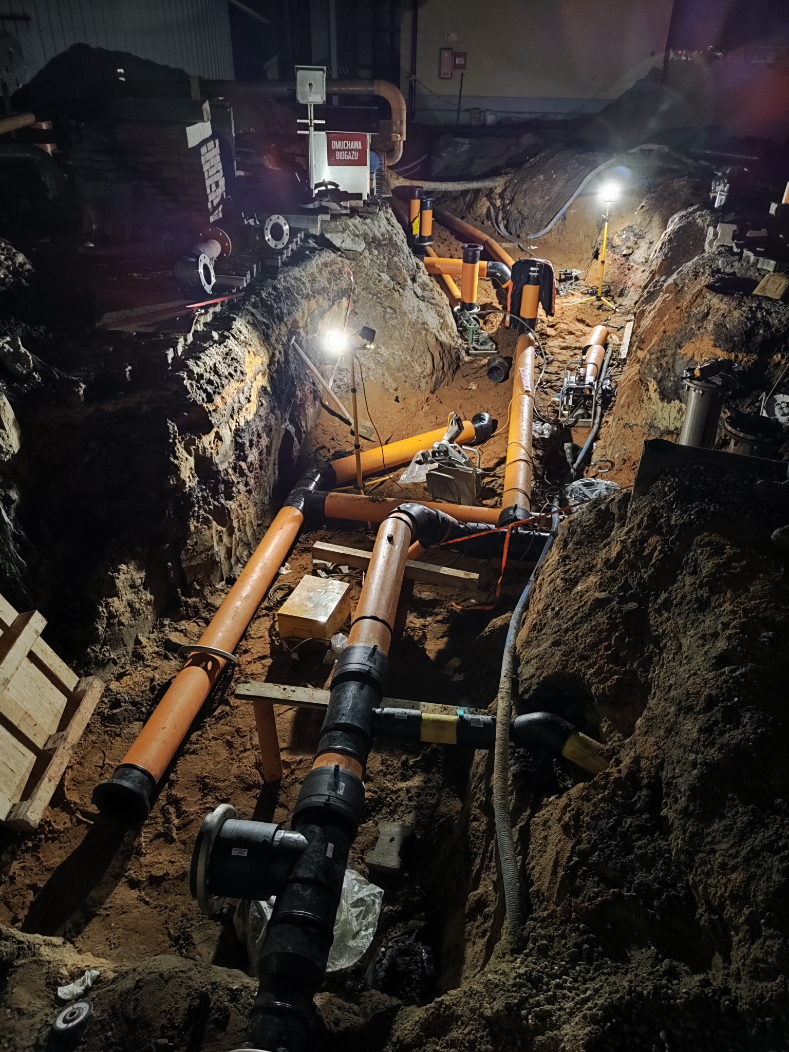 Wykop z ułożonymi rurami wodno-kanalizacyjnymi i gazowymi, widoczne złącza, oświetlenie budowlane i tablica informacyjna w tle.