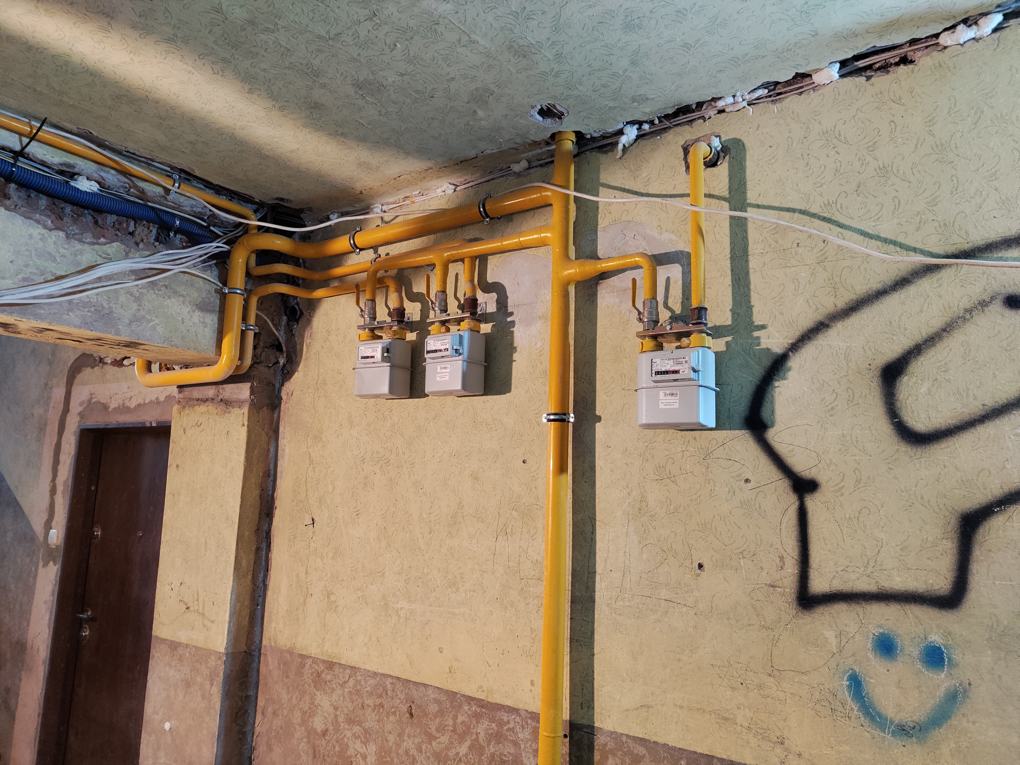 Żółte rury gazowe prowadzone po suficie i ścianie wewnątrz budynku, z zamontowanymi trzema licznikami gazowymi, widoczne niedociągnięcia w wykończeniu, graffiti na ścianie.