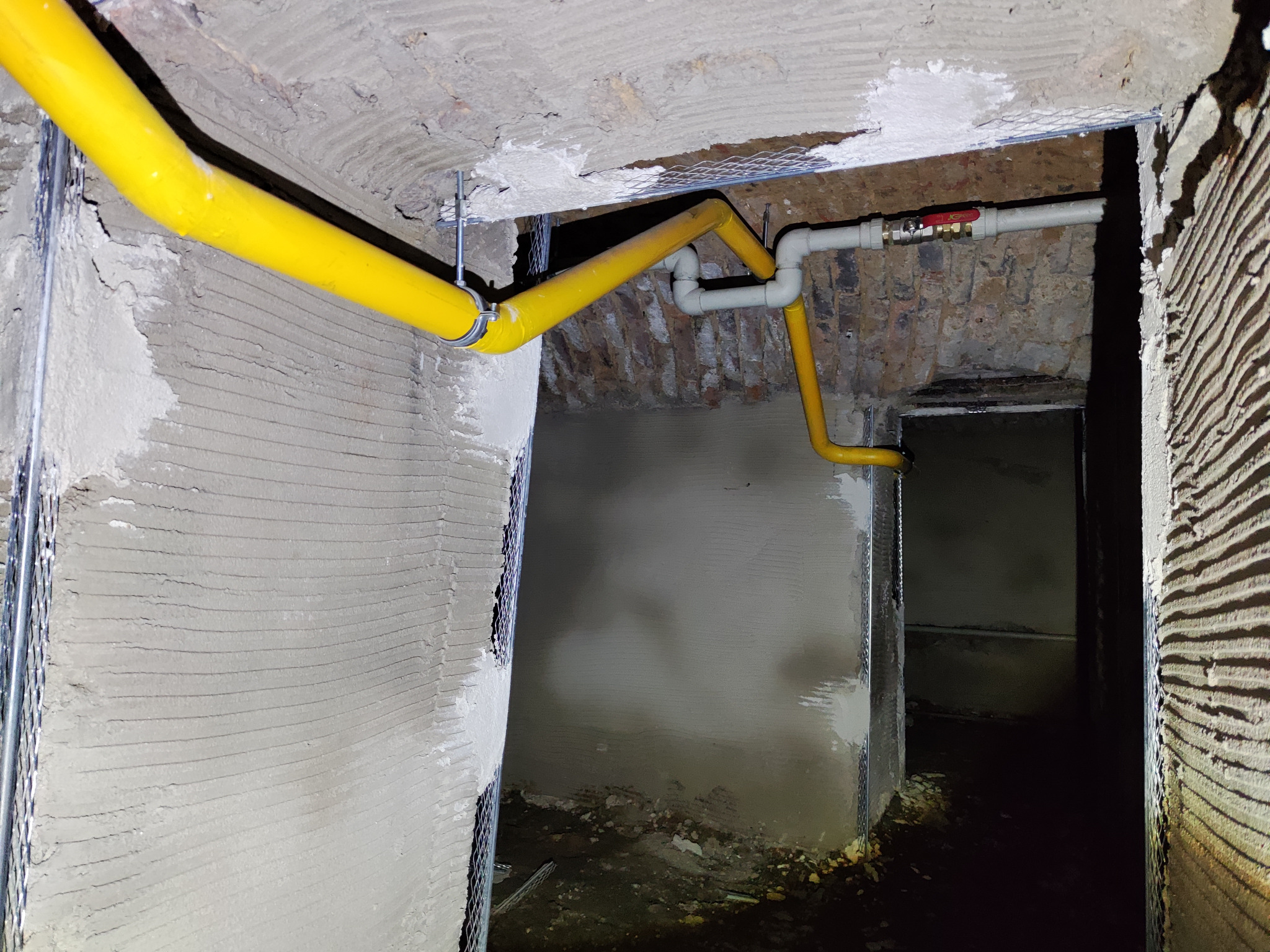 Żółta rura gazowa zamontowana pod sufitem w surowym pomieszczeniu, widoczne fragmenty ścian z siatką i świeżym tynkiem oraz zawór odcinający na rurze.