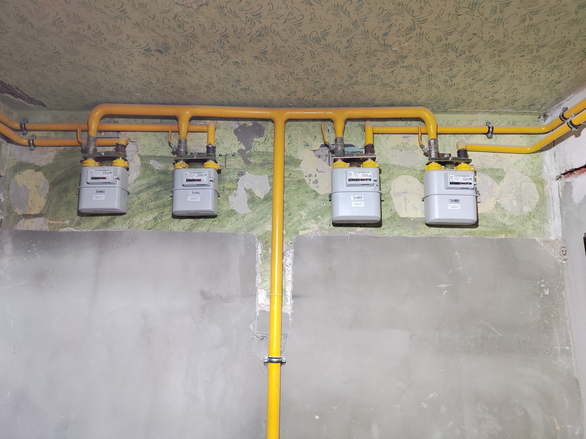 Żółta instalacja gazowa z czterema licznikami na tle ściany z resztkami tapety i surowym tynkiem.