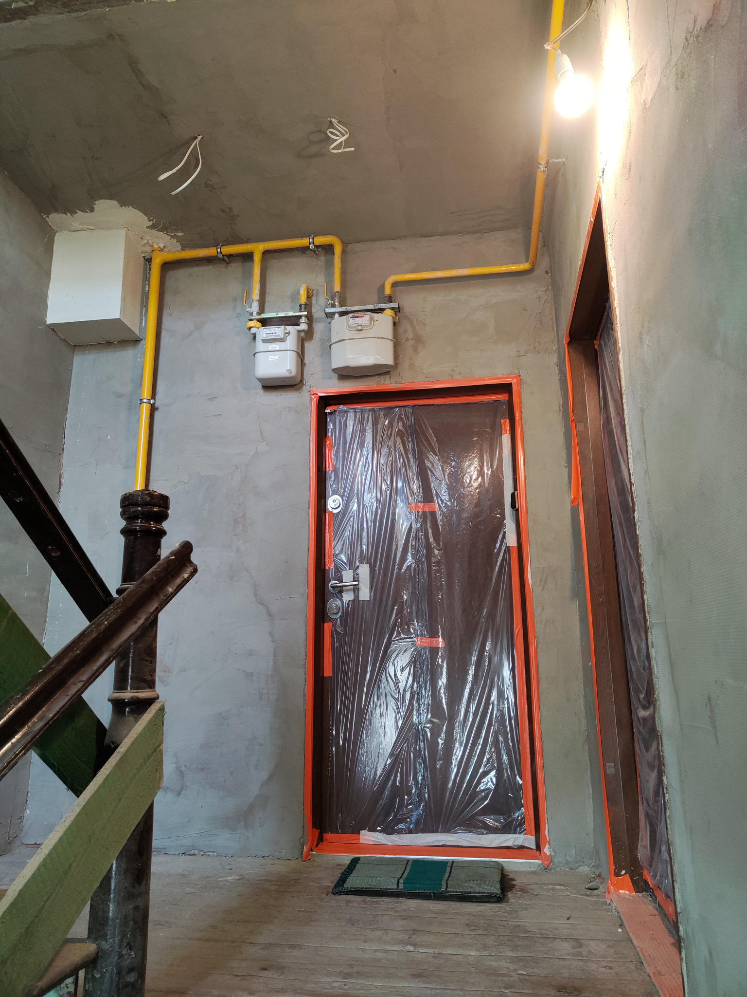 Wewnętrzna instalacja gazowa z dwoma licznikami, żółte rury prowadzone wzdłuż szarej ściany, zabezpieczone folią drzwi wejściowe, widoczne elementy klatki schodowej, surowy stan wykończenia wnętrza.