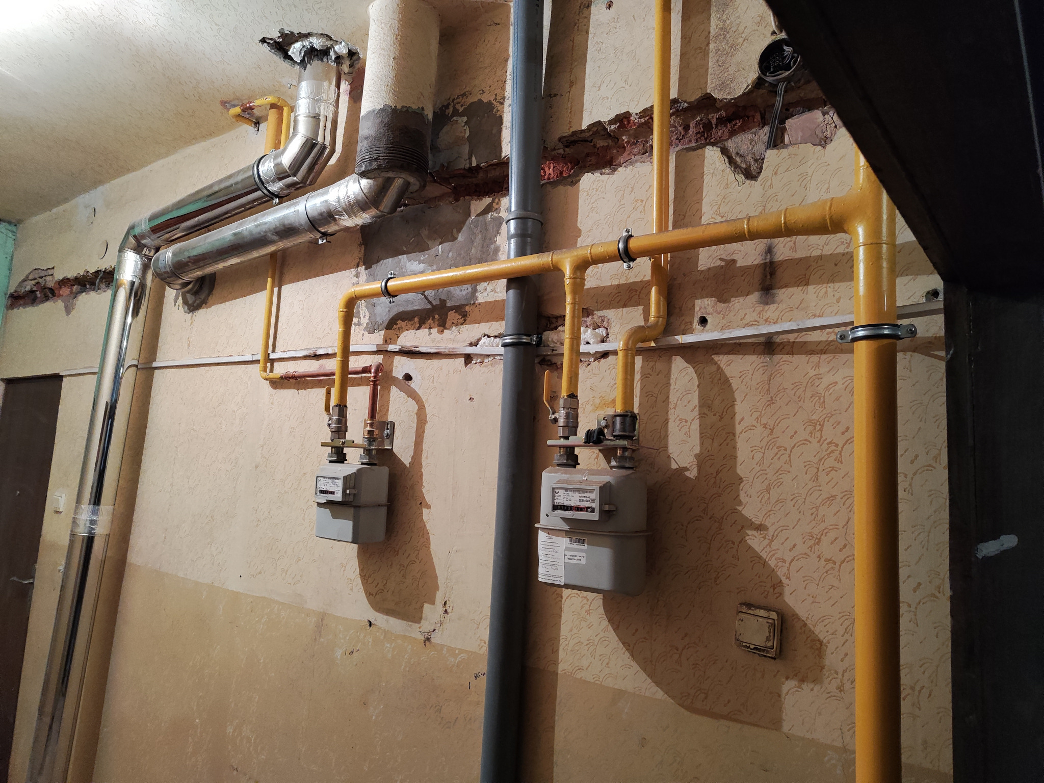 Żółte rury gazowe i srebrne rury wentylacyjne zamontowane na ścianie z widocznymi śladami po remoncie i dwoma szarymi gazomierzami, widoczne również fragmenty instalacji wodno-kanalizacyjnej.
