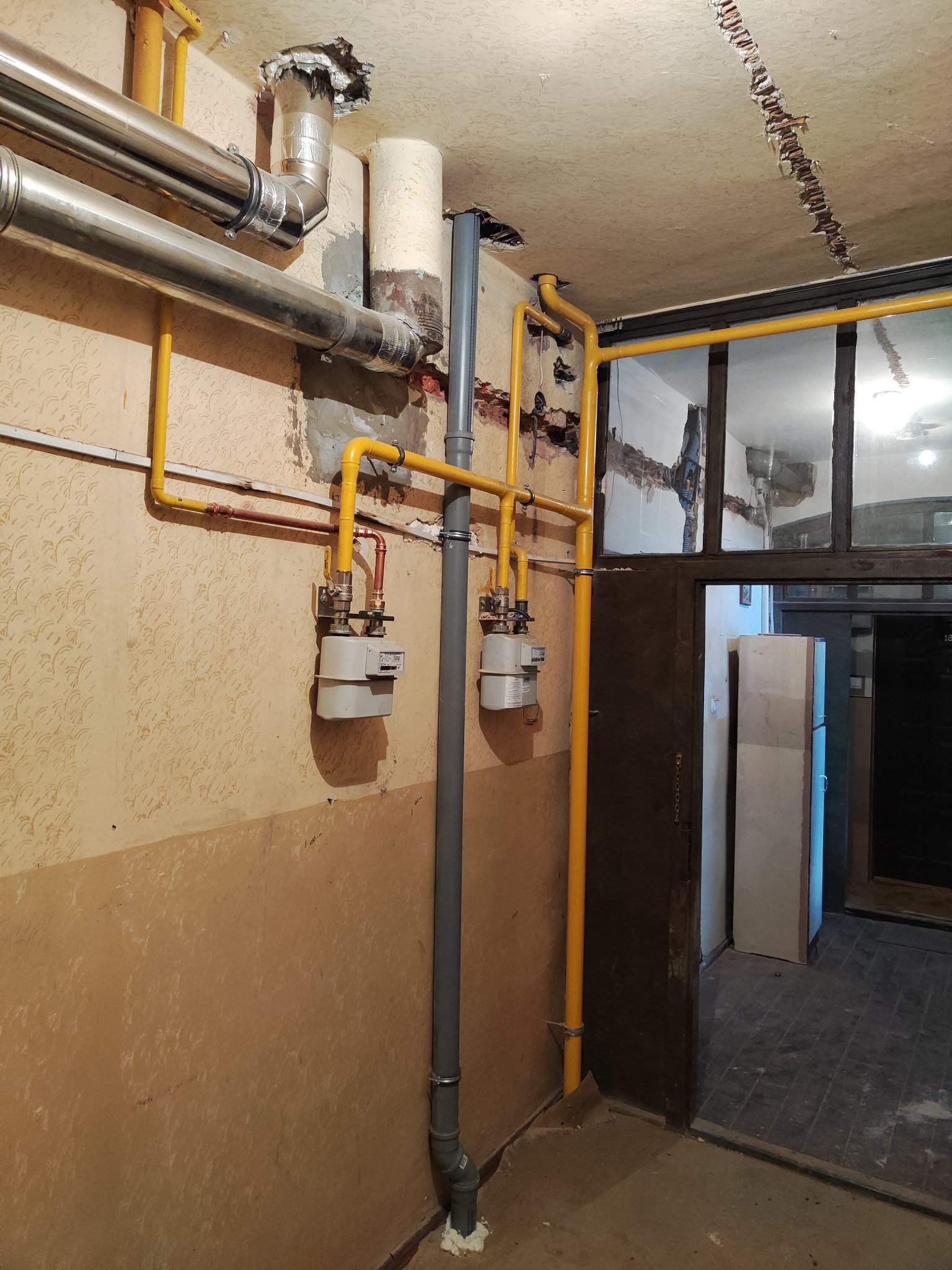 Dwa gazomierze zamontowane na ścianie z żółtymi rurami gazowymi, widoczne również rury wentylacyjne, ściany w jasnych kolorach z widocznymi śladami po remoncie.