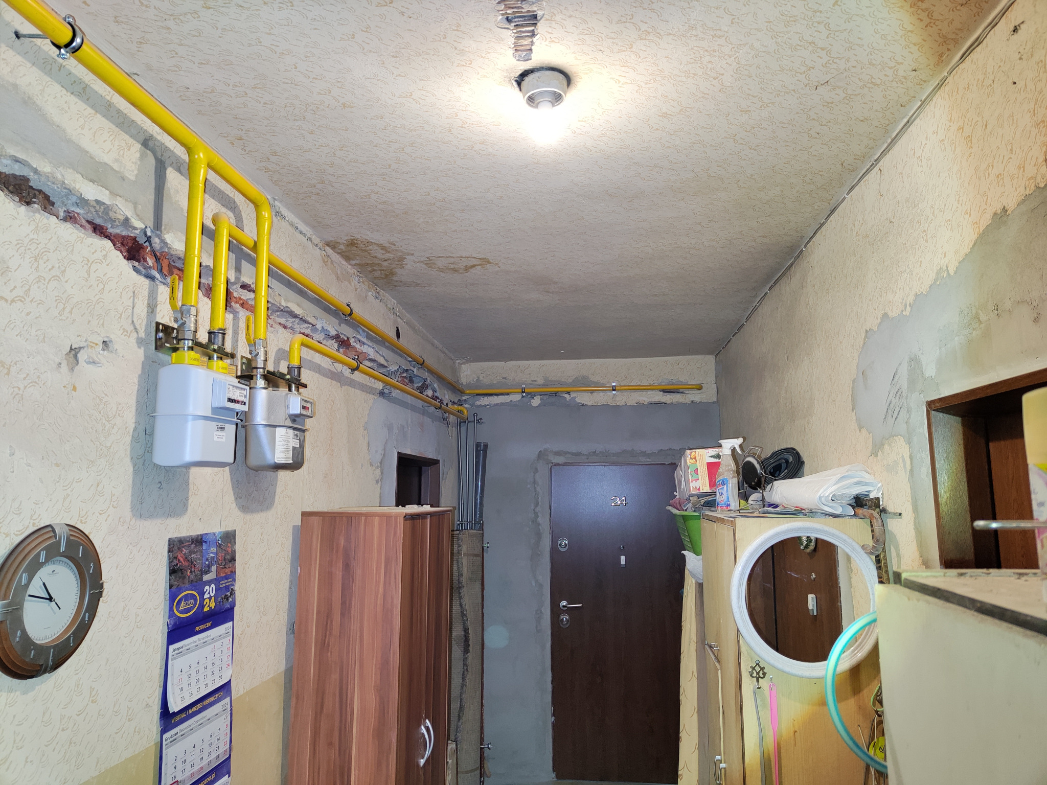Wnętrze korytarza z widoczną instalacją gazową: żółte rury i trzy liczniki gazowe na ścianie, obok zegar i kalendarz na rok 2024, ściany w trakcie remontu, sufit z widocznymi plamami, drzwi...