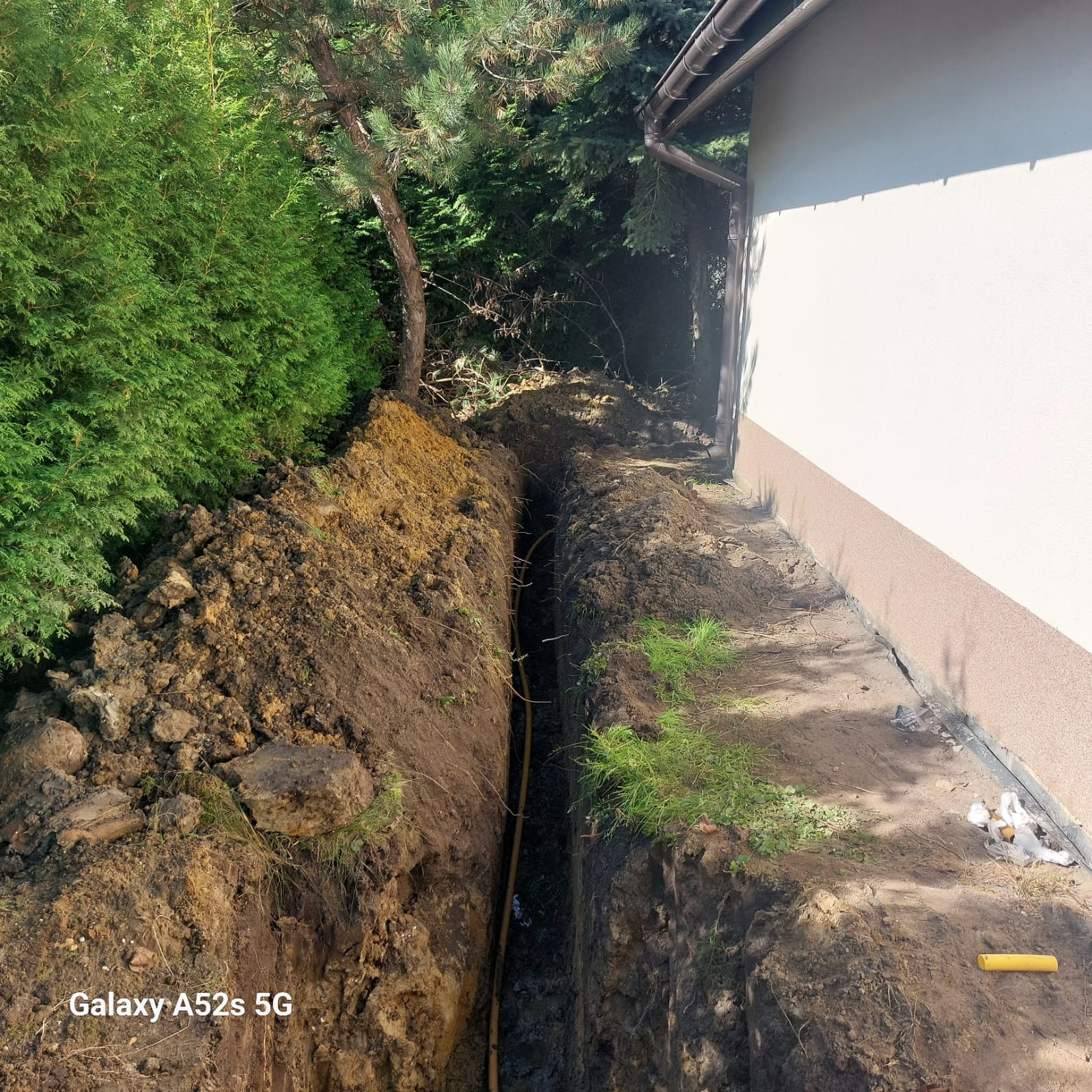 Wykopany rów w ziemi obok budynku z widoczną żółtą rurą w środku, prawdopodobnie gazową, z kupą ziemi obok i roślinnością w tle.