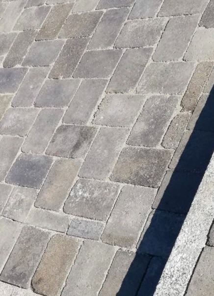 Ułożona kostka brukowa w odcieniach szarości, ułożona w romby. Częściowo widoczny element obrzeża. Widok z góry, oświetlenie dzienne.