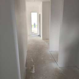 PRO-TYNK KINGA PAWLAK - Korytarz w stanie surowym po wstępnym malowaniu ścian na biało, widoczne wystające przewody elektryczne z sufitu, betonowa podłoga z resztkami zaprawy, oraz osoba w oddali za przeszklonymi drzwiami.