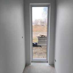 PRO-TYNK KINGA PAWLAK - Wnętrze pokoju po świeżym malowaniu ścian na biało, widoczne gładkie powierzchnie, betonowa posadzka i okno z widokiem na stos płyt na zewnątrz.