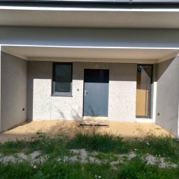 PRO-TYNK KINGA PAWLAK - Nowoczesna elewacja domu z szarą, gładką strukturą, widoczne okno i drzwi wejściowe, teren przed budynkiem częściowo pokryty piaskiem i rosnącą trawą.