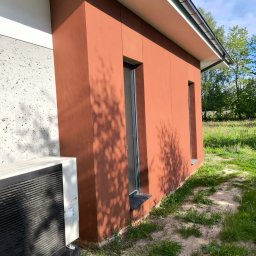 PRO-TYNK KINGA PAWLAK - Fragment elewacji domu z oknami w kolorze terakoty, z widocznym klimatyzatorem na fundamencie i cieniem drzew na ścianie.