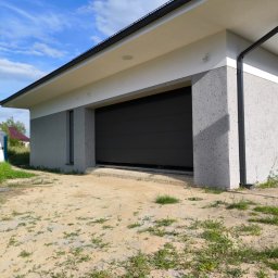 PRO-TYNK KINGA PAWLAK - Garaż z szarą, segmentową bramą wjazdową i elewacją w odcieniach szarości i bieli, z widocznym fragmentem dachu z czarną rynną, na tle błękitnego nieba z chmurami.