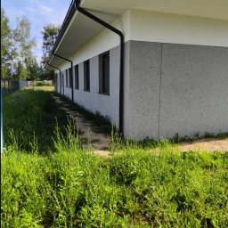 PRO-TYNK KINGA PAWLAK - Nowoczesny budynek z szarą elewacją typu baranek i białym pasem pod dachem, widok z poziomu trawy