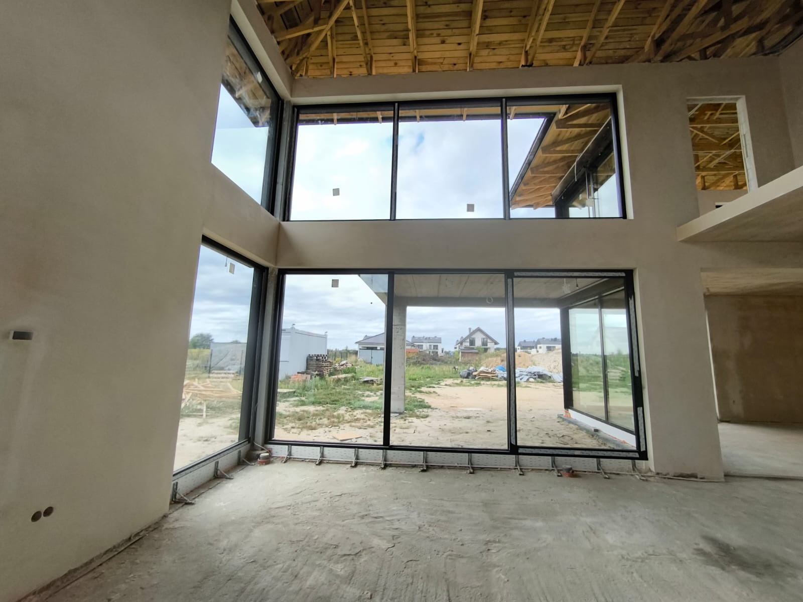 Wnętrze domu w stanie surowym otwartym z dużymi oknami aluminiowymi w czarnych ramach, widok na zewnątrz na budowę i domy w tle, surowy strop z drewnianych belek.