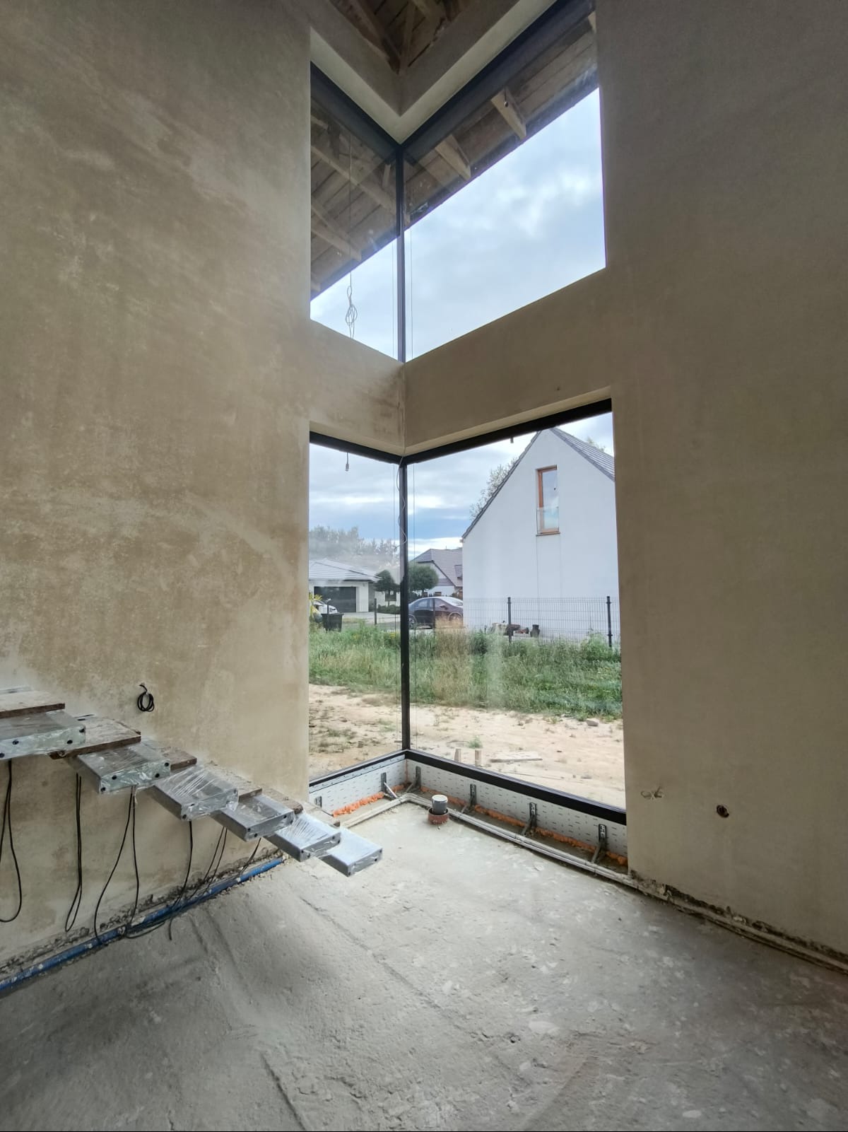 Surowe ściany w nowym domu z dużymi oknami, widok na zewnątrz na dom i zieleń, tymczasowe schody prowadzące na górę.