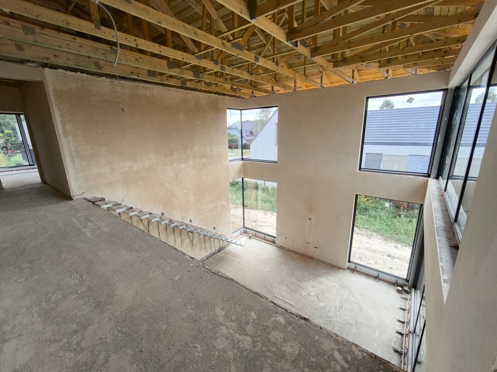 Surowy stan wnętrza domu z wysokimi oknami, ściany po tynkowaniu, widoczne elementy instalacji elektrycznej, drewniana konstrukcja dachu, betonowa podłoga w trakcie prac wykończeniowych.