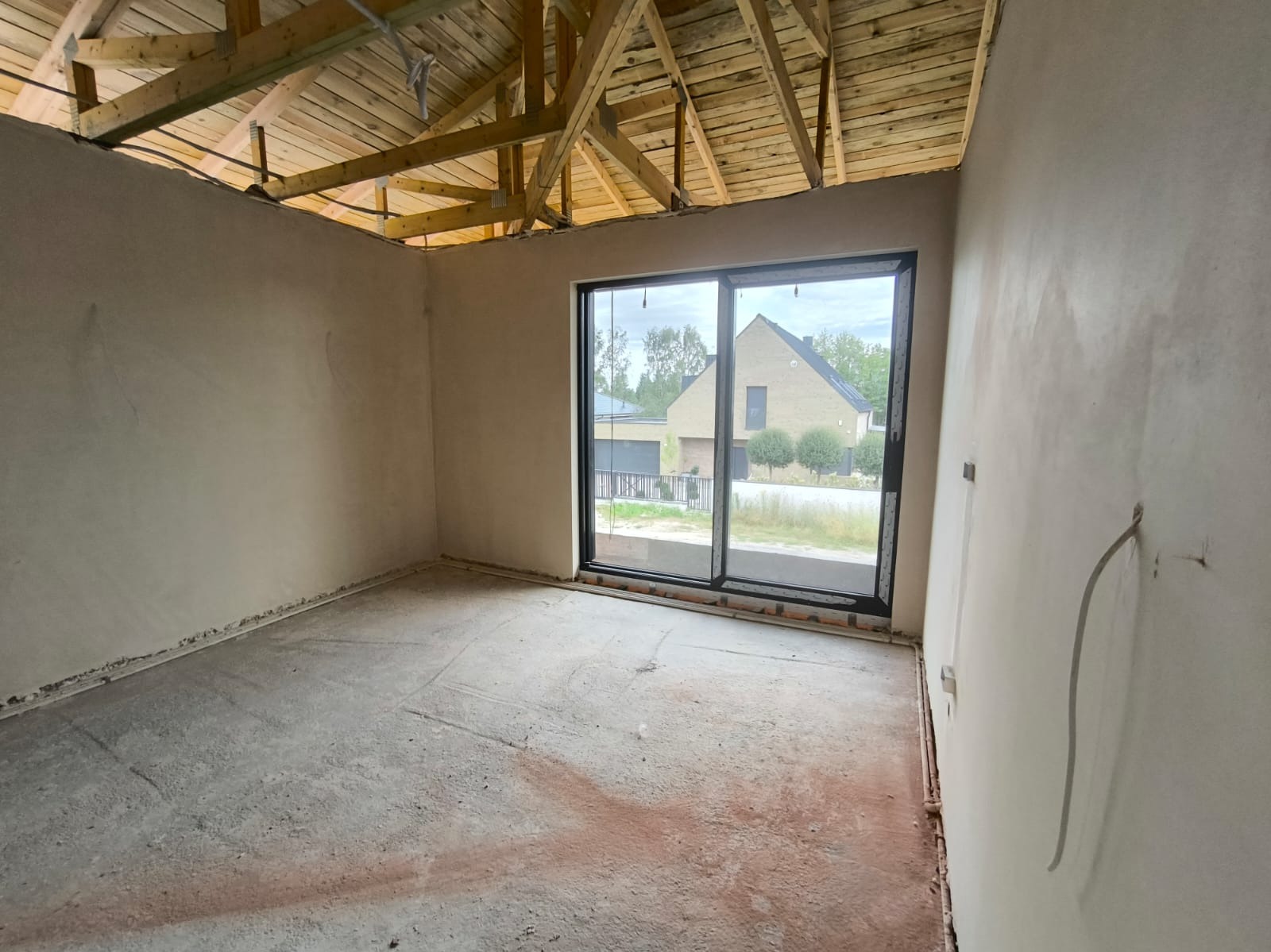 Wnętrze w stanie deweloperskim z surowymi ścianami po tynkowaniu, widoczna drewniana konstrukcja dachu i duże okno przesuwne z widokiem na dom sąsiada.