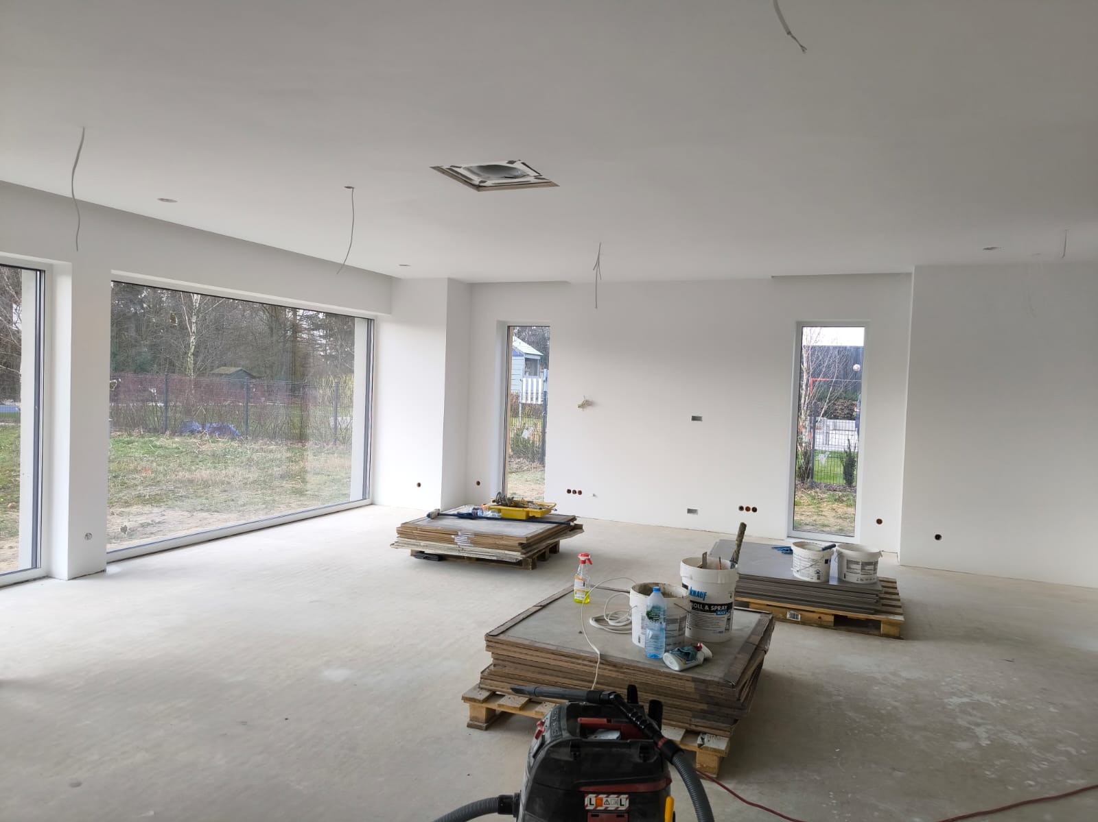 Wnętrze nowo wykończonego, jasnego pomieszczenia z dużymi oknami. Widoczne przygotowane materiały budowlane na paletach i odkurzacz przemysłowy w rogu. Ściany i sufit świeżo pomalowane na biało,...