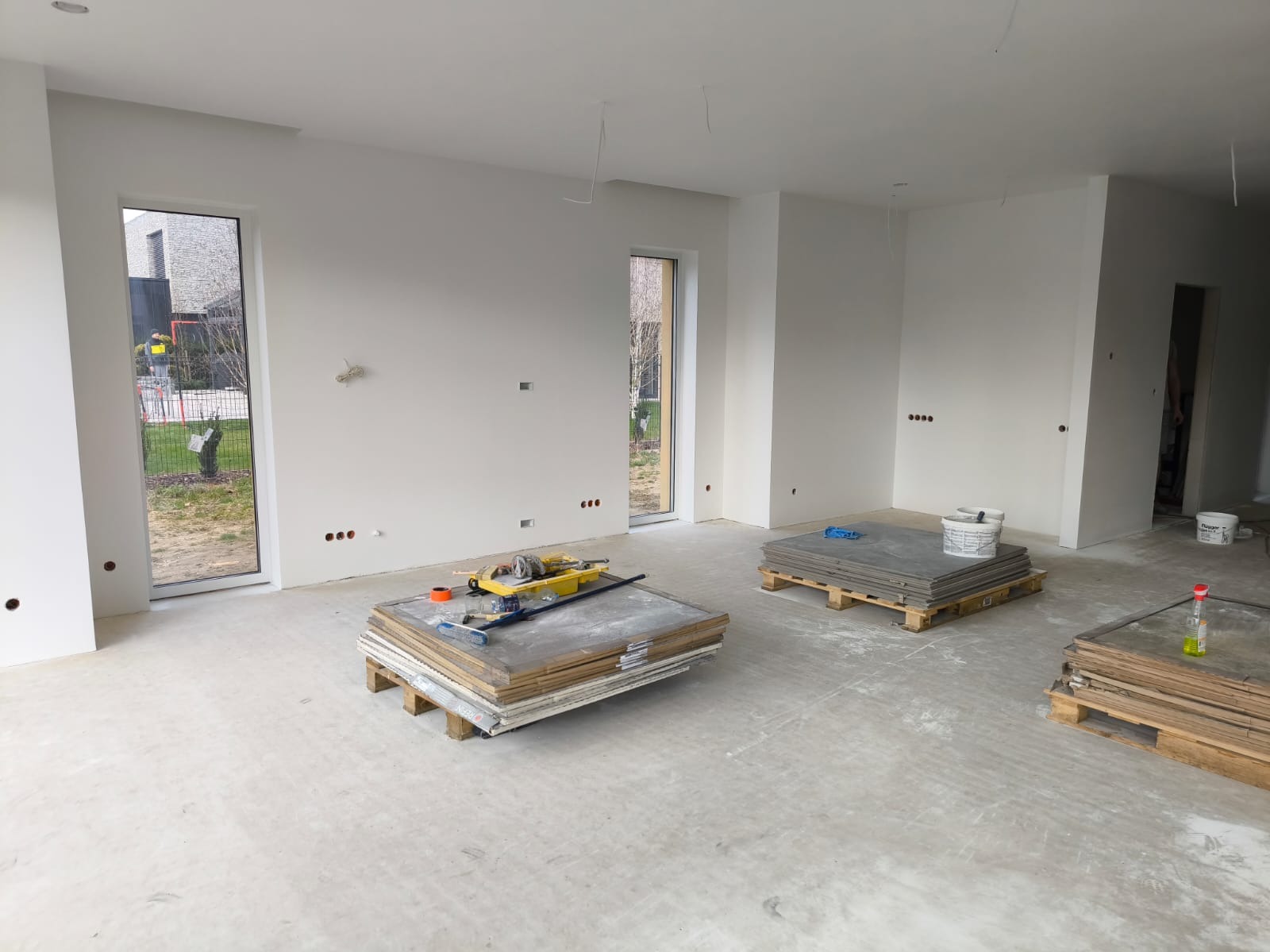 Wnętrze w trakcie prac wykończeniowych, świeżo pomalowane ściany na biało, okna z widokiem na zewnątrz, na podłodze palety z płytami, widoczna szlifierka do gipsu i akcesoria malarskie.