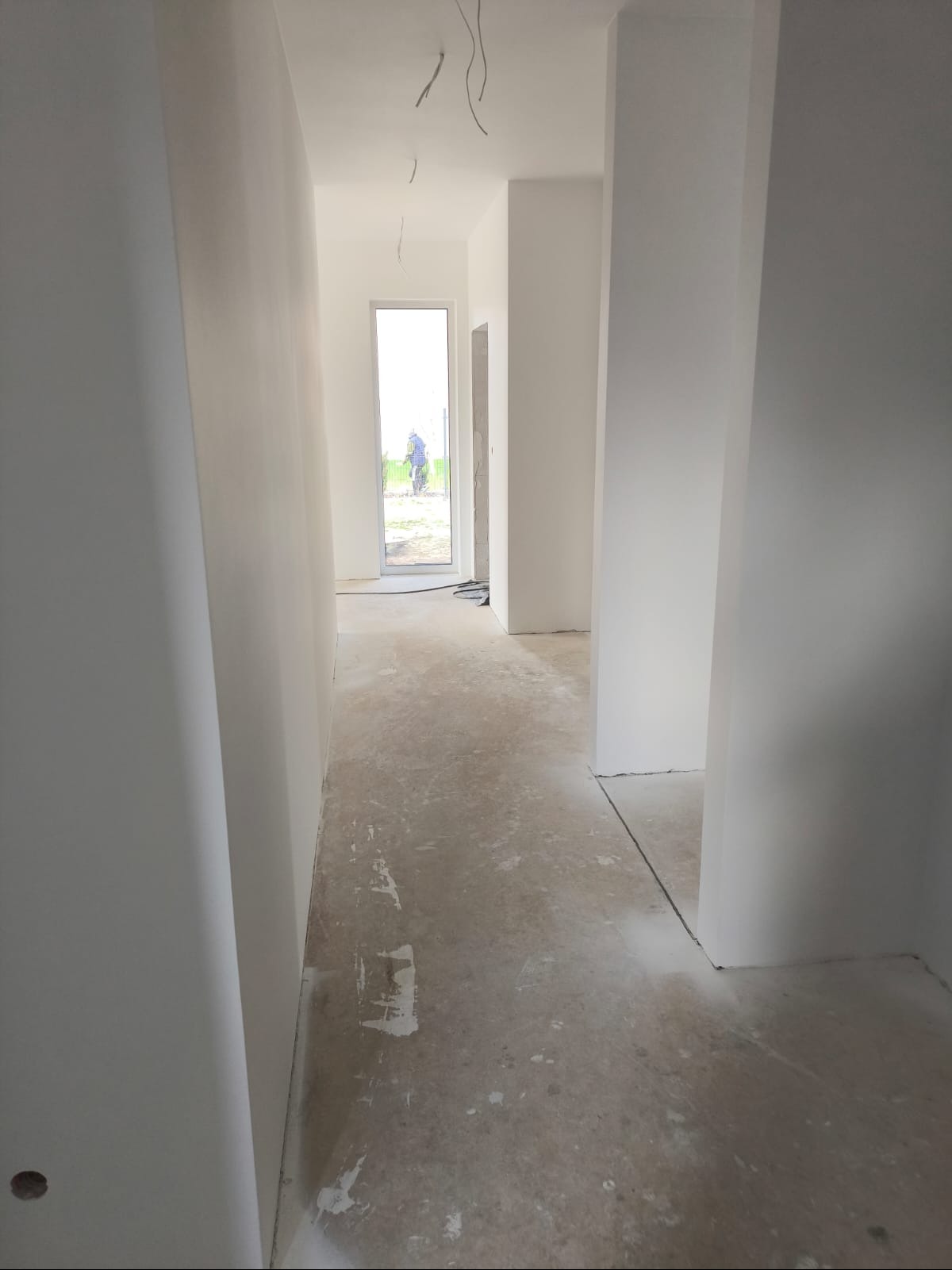 Korytarz w stanie surowym po wstępnym malowaniu ścian na biało, widoczne wystające przewody elektryczne z sufitu, betonowa podłoga z resztkami zaprawy, oraz osoba w oddali za przeszklonymi drzwiami.