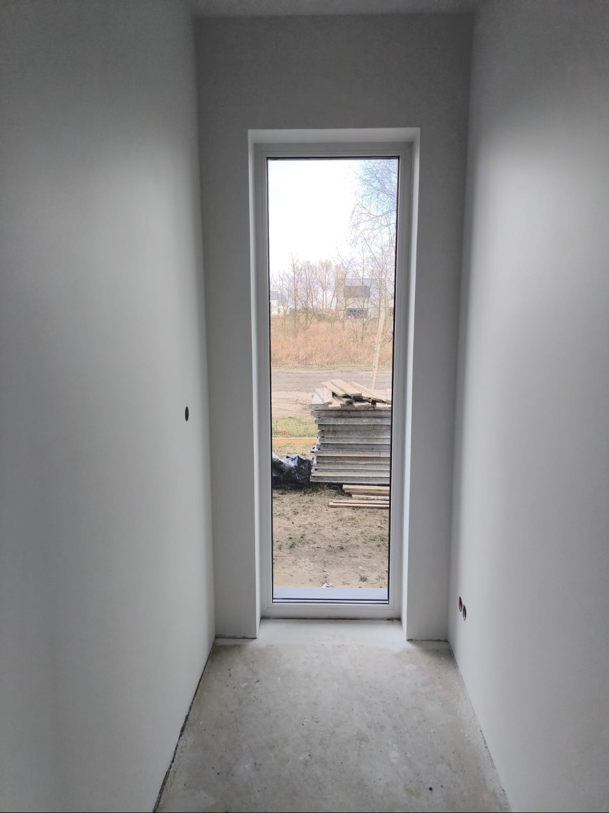 Wnętrze pokoju po świeżym malowaniu ścian na biało, widoczne gładkie powierzchnie, betonowa posadzka i okno z widokiem na stos płyt na zewnątrz.