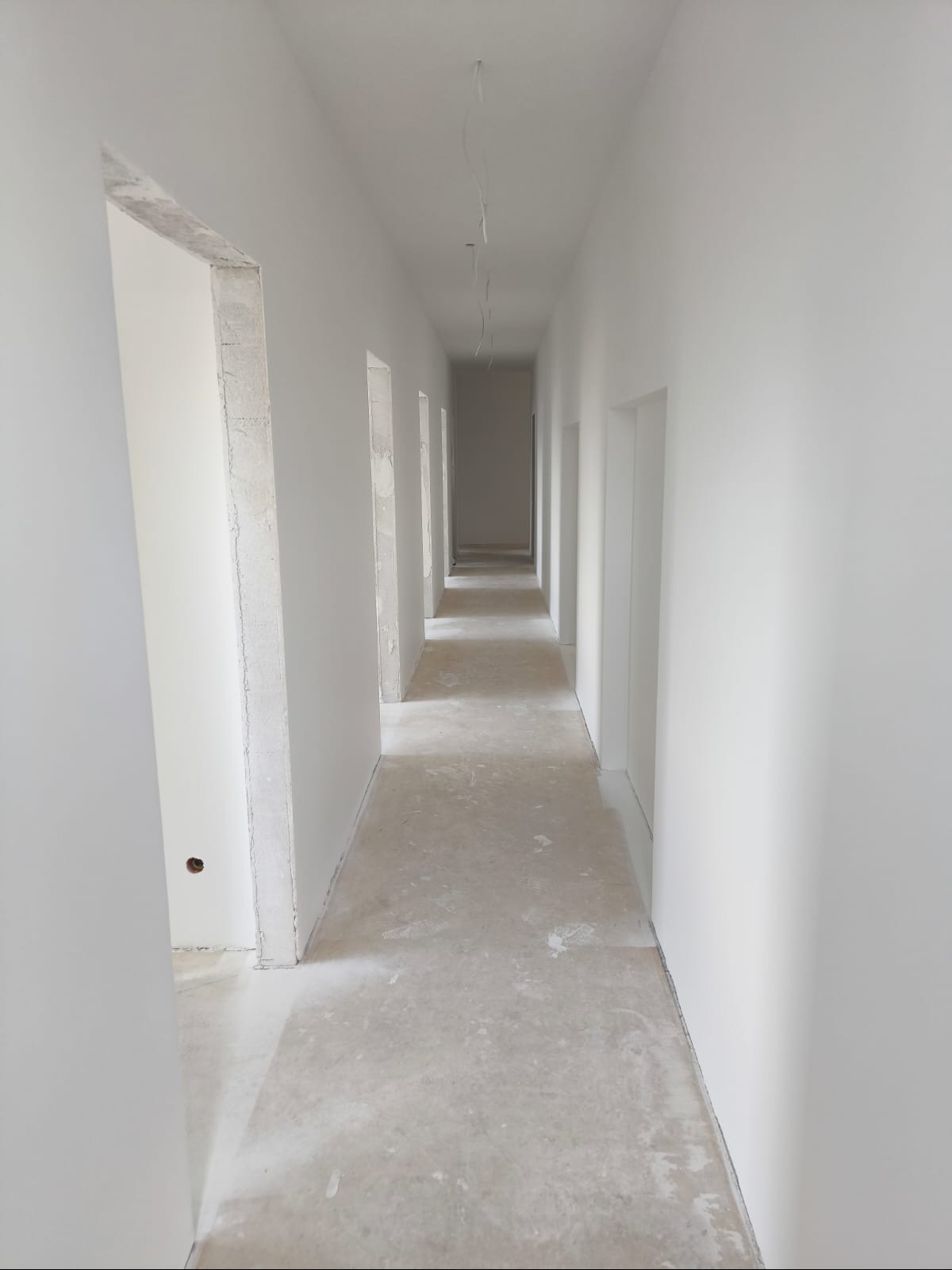 Długi korytarz w trakcie prac wykończeniowych, świeżo pomalowane ściany na biało, surowy beton na podłodze, widoczne wystające kable ze stropu, ościeżnice drzwiowe w stanie surowym.