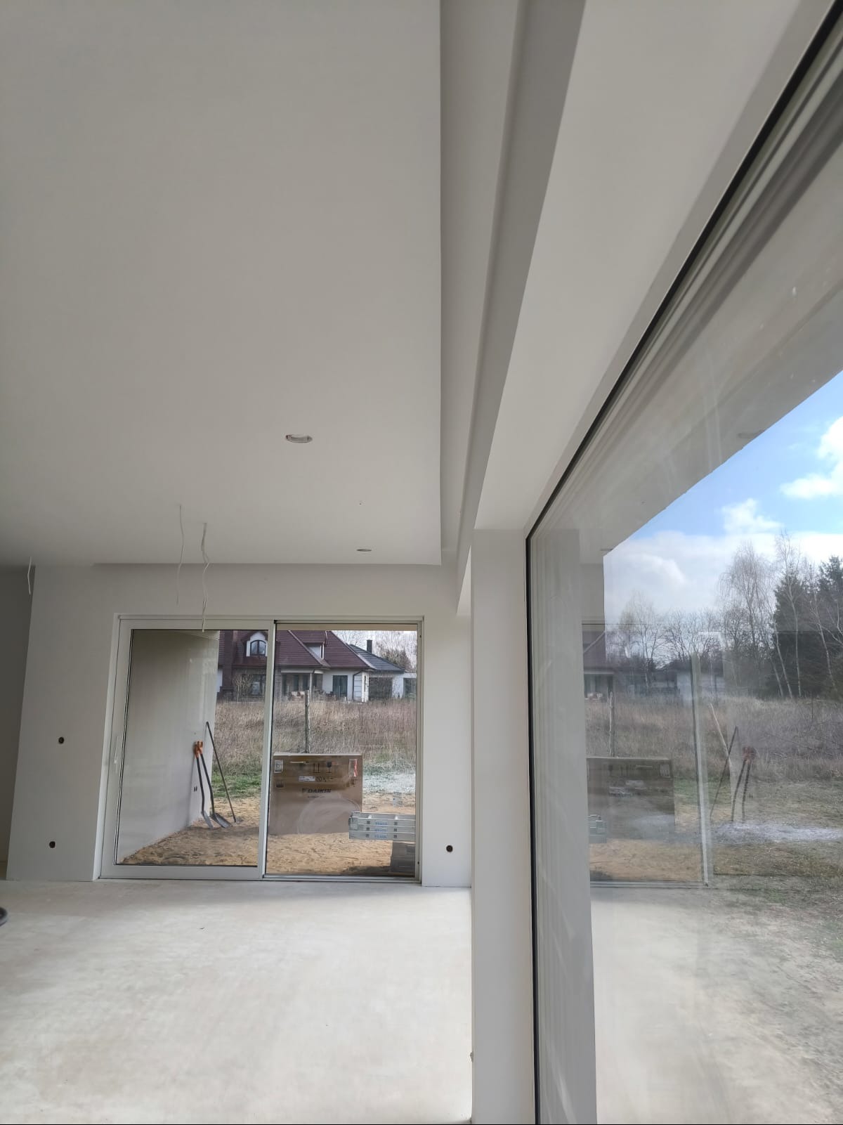 Wnętrze w stanie deweloperskim z widocznymi gładziami na ścianach i suficie, duże okna przesuwne z widokiem na domy w tle, przygotowane do dalszych prac wykończeniowych.