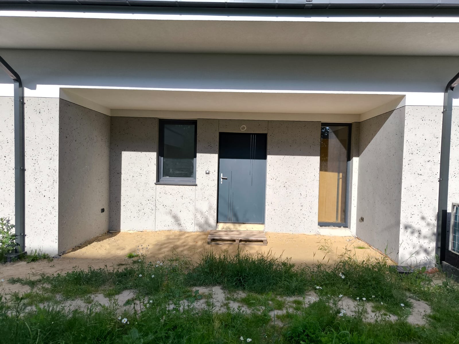 Nowoczesna elewacja domu z szarą, gładką strukturą, widoczne okno i drzwi wejściowe, teren przed budynkiem częściowo pokryty piaskiem i rosnącą trawą.
