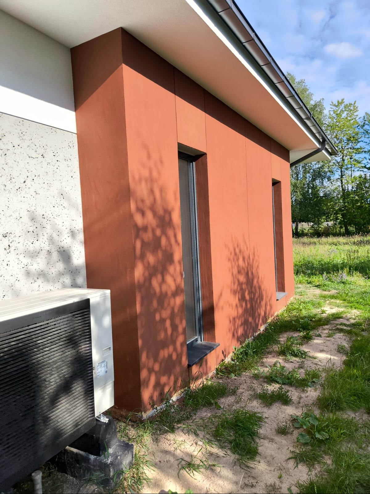Fragment elewacji domu z oknami w kolorze terakoty, z widocznym klimatyzatorem na fundamencie i cieniem drzew na ścianie.