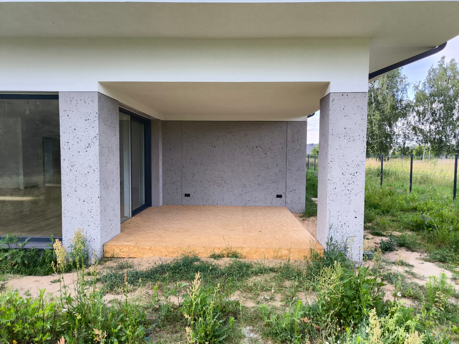 Zadaszony taras w surowym stylu z płytą OSB jako tymczasową podłogą, szare ściany z efektem betonu architektonicznego, widok na zarośnięty ogród za czarnym ogrodzeniem.