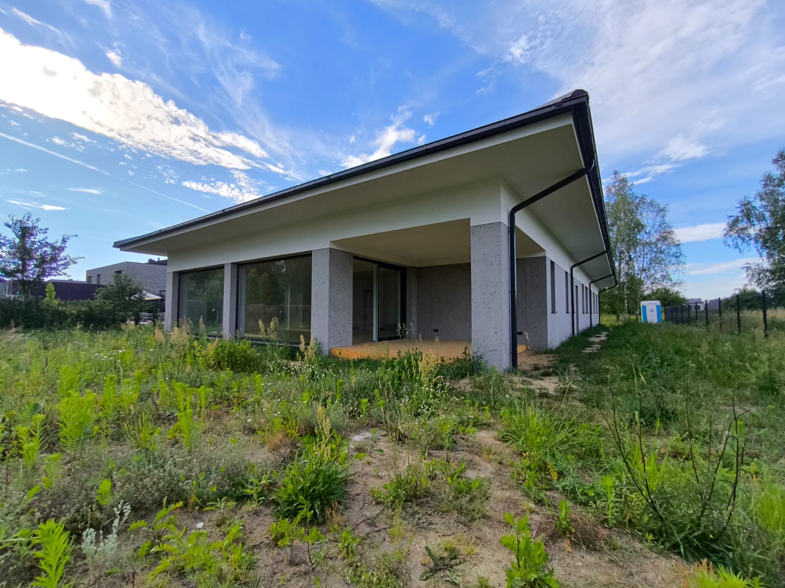 Nowoczesny parterowy dom z płaskim dachem, szare betonowe kolumny, duże okna, widok z zarośniętego trawnika, błękitne niebo z chmurami