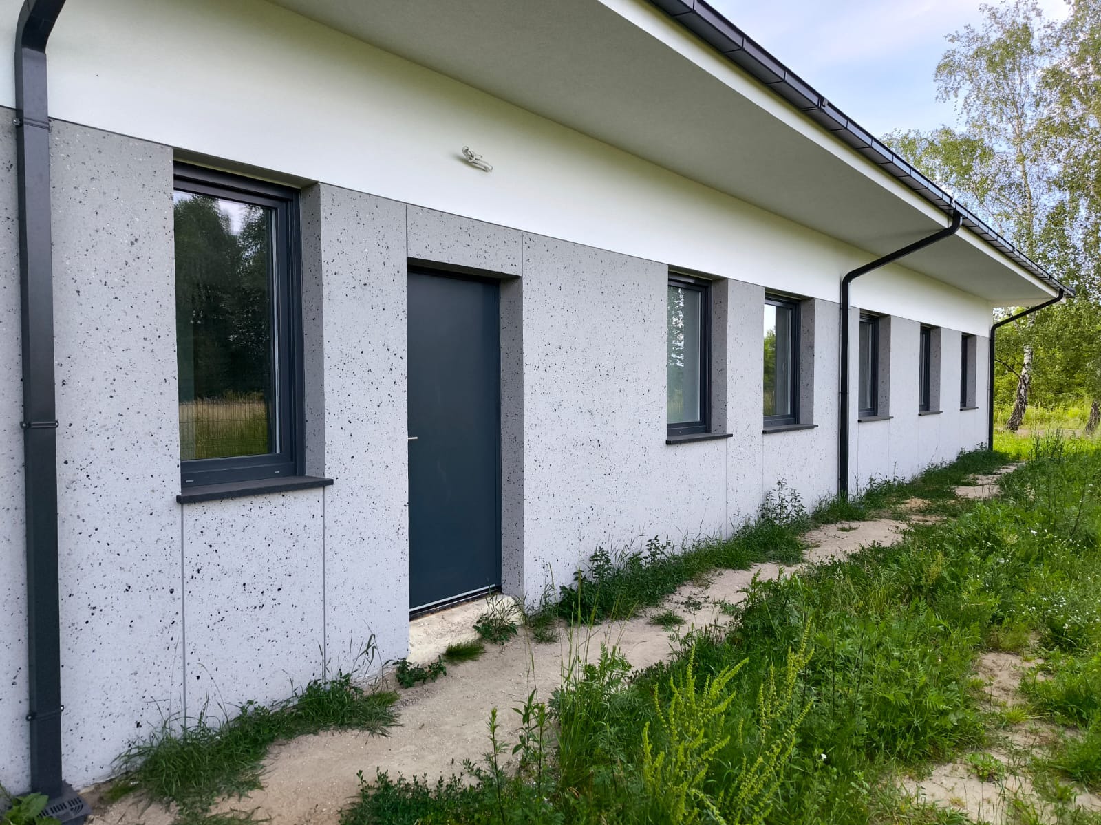 Elewacja budynku wykończona panelami imitującymi lastryko, antracytowa stolarka okienna i drzwiowa, widoczne rynny spustowe.