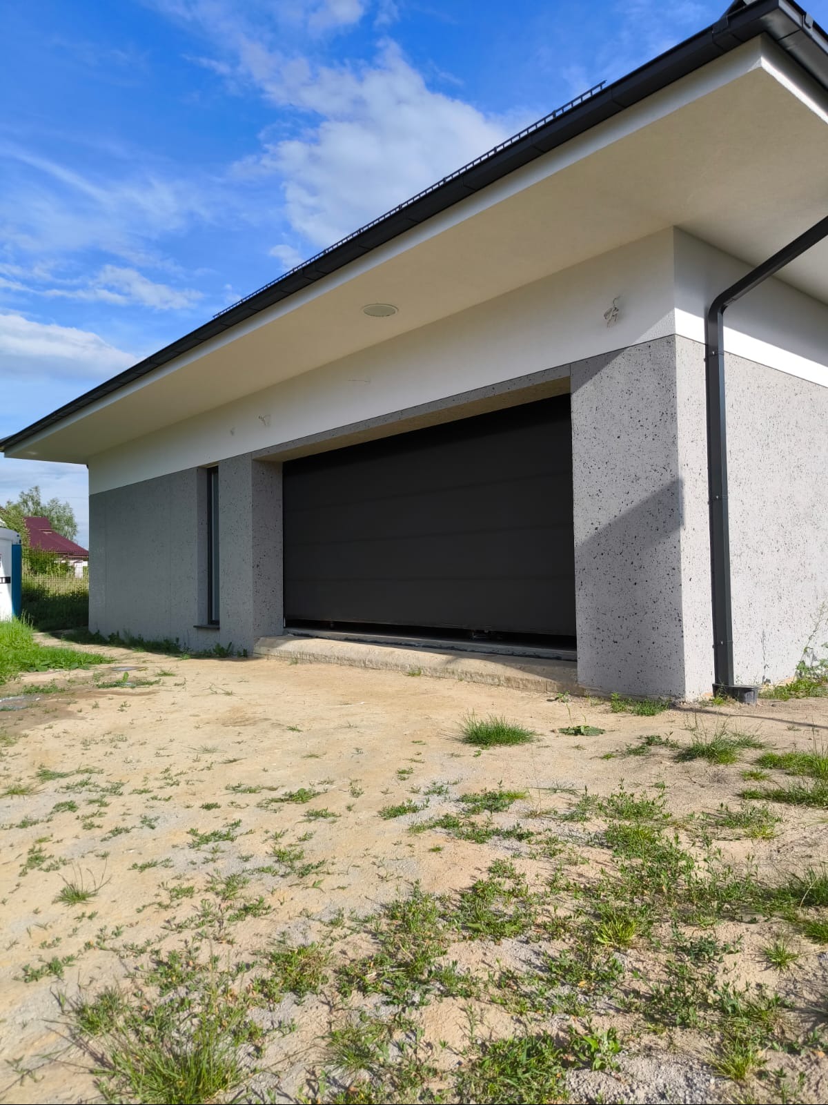 Garaż z szarą, segmentową bramą wjazdową i elewacją w odcieniach szarości i bieli, z widocznym fragmentem dachu z czarną rynną, na tle błękitnego nieba z chmurami.